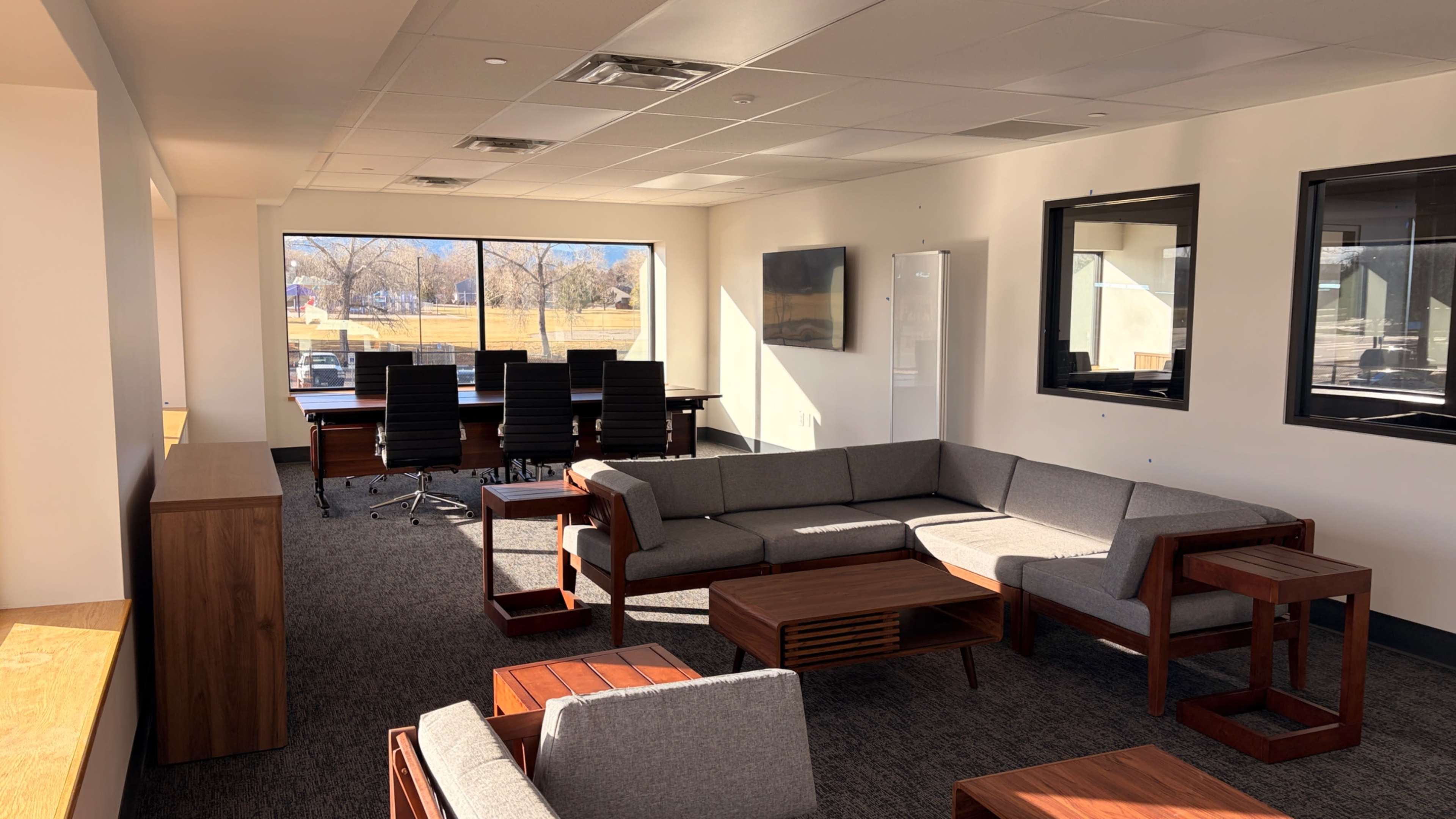 The image shows a modern office space featuring a lounge area with gray sofas and wooden tables, alongside a conference table in a well-lit room with large windows.