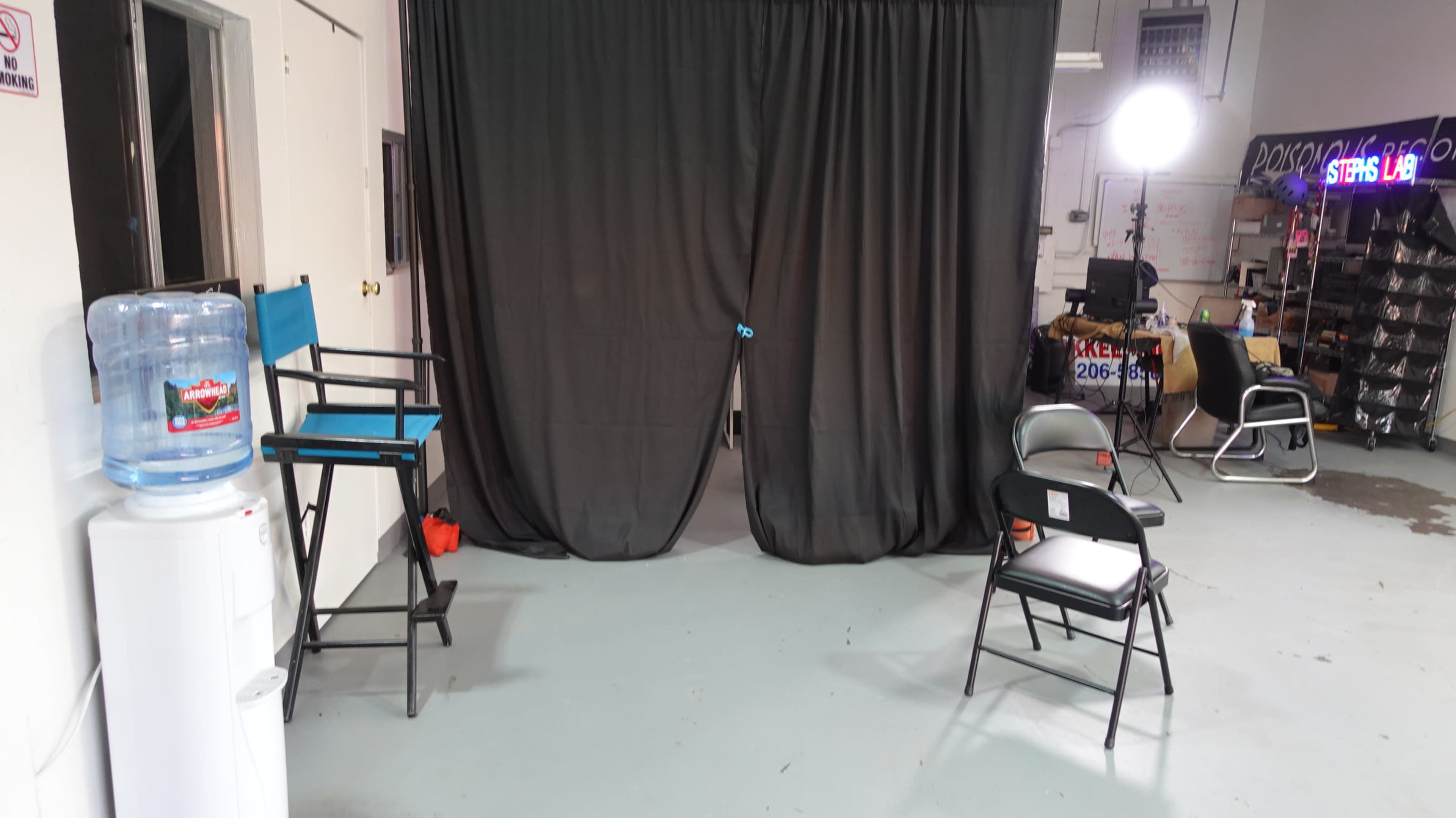 The image shows a small studio or rehearsal space with a black curtain, two chairs, and a large water cooler against a white wall.