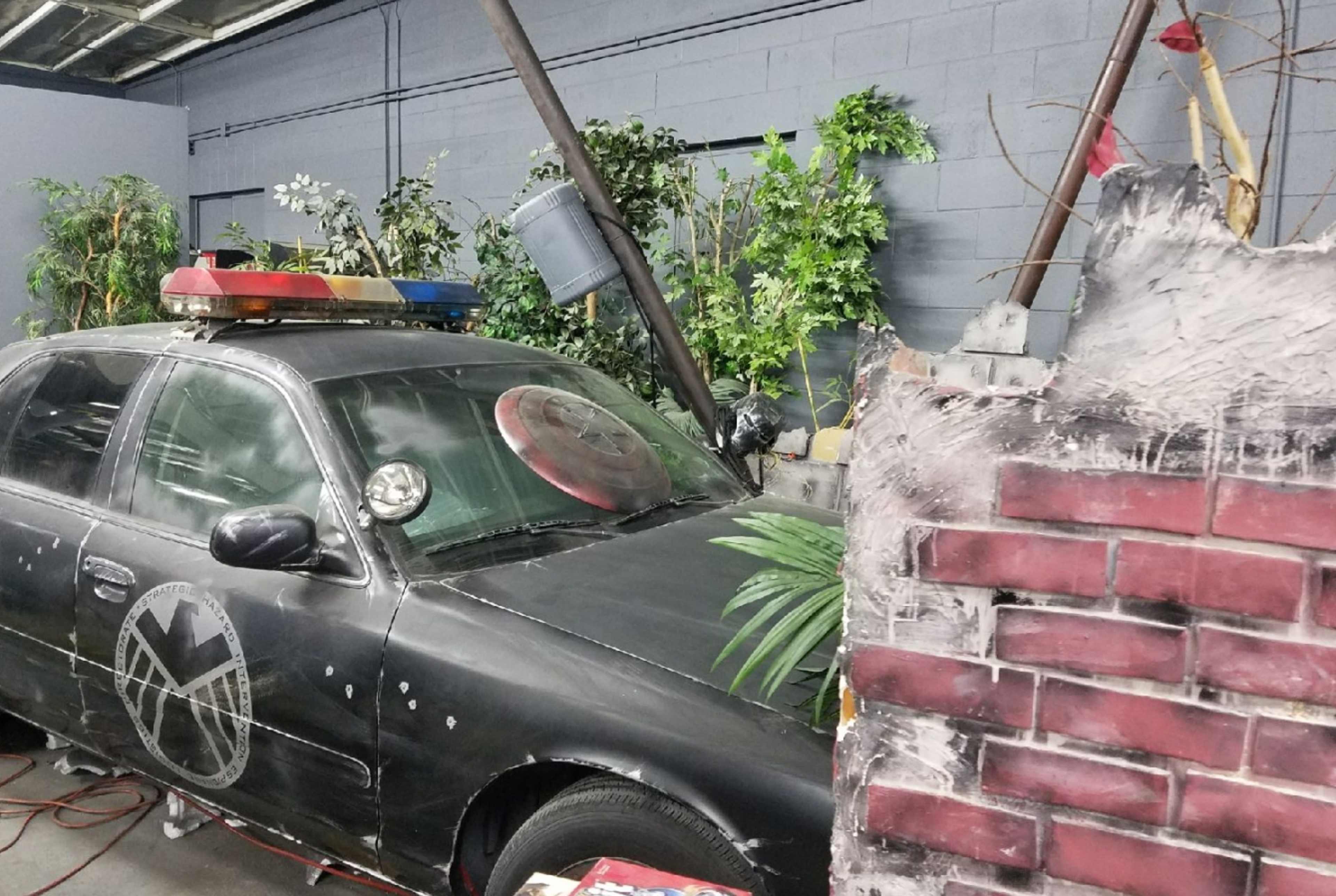 A black car with police lights parked near a wall partially covered in brick-patterned material and surrounded by various plants and branches.