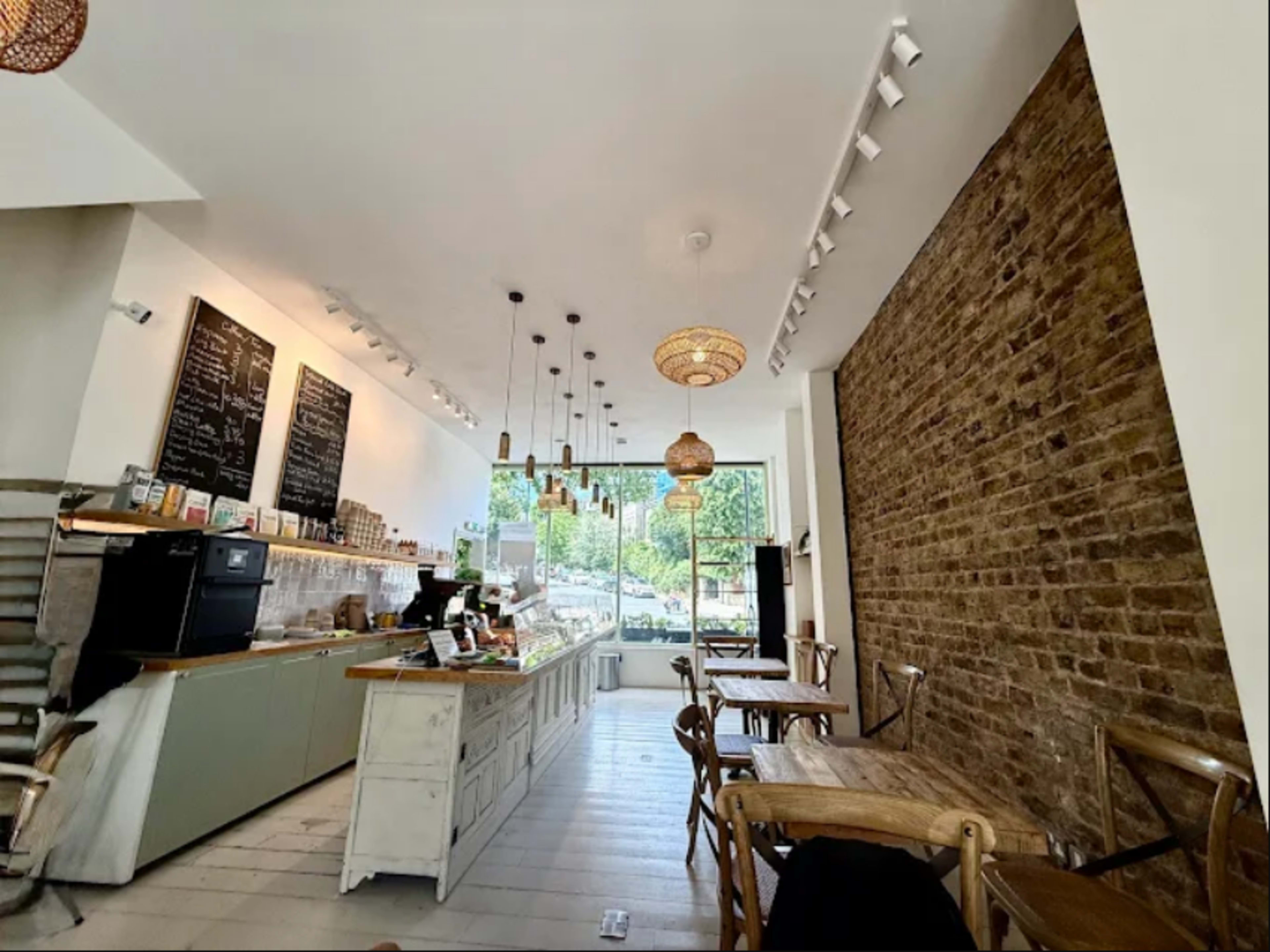 The image shows a modern café with a light-colored interior, featuring a service counter, wooden tables, and a large window providing natural light.