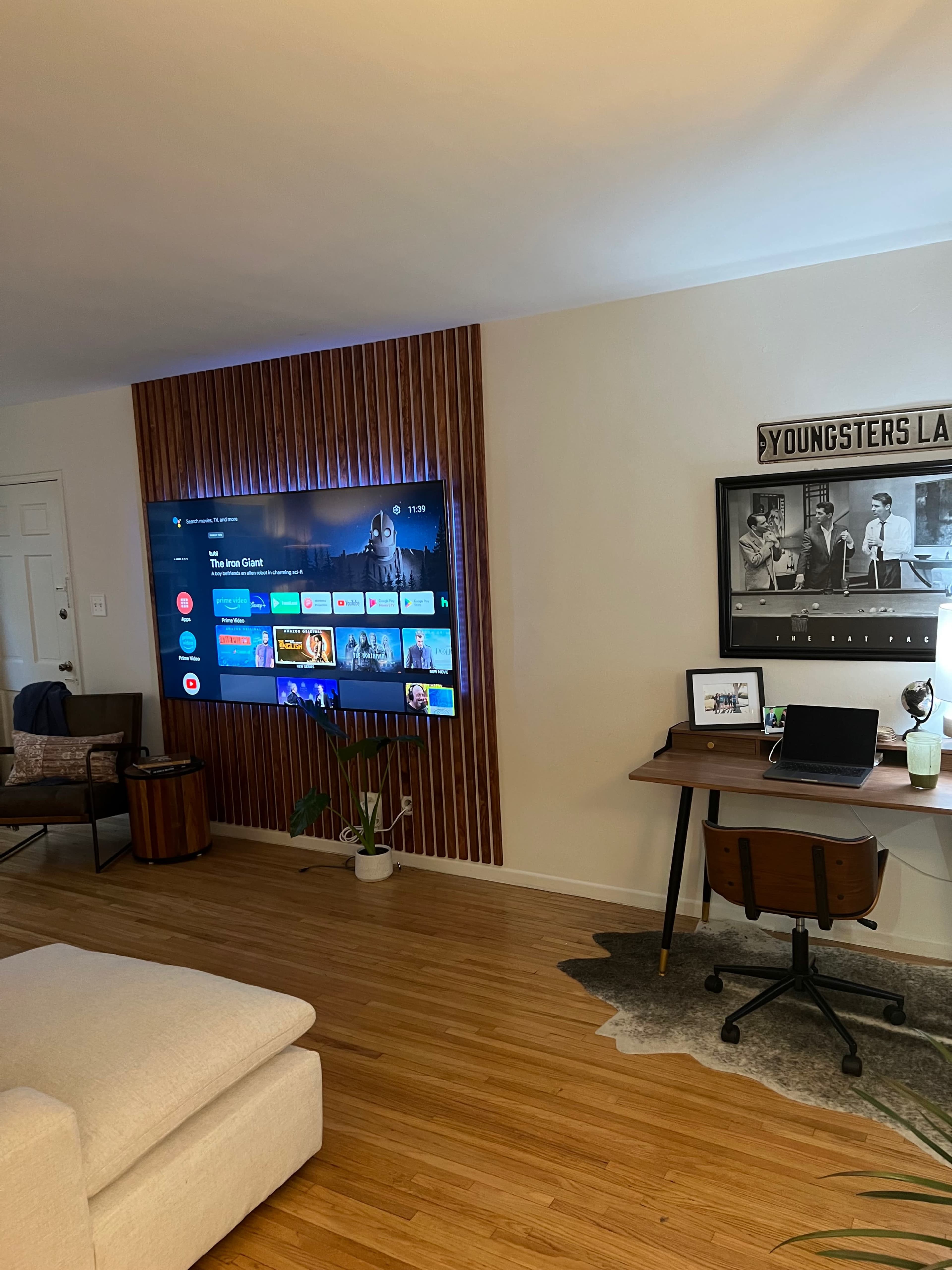 A modern living room features a wall-mounted television and a desk area with a chair, surrounded by wooden accents and a rug on the floor.