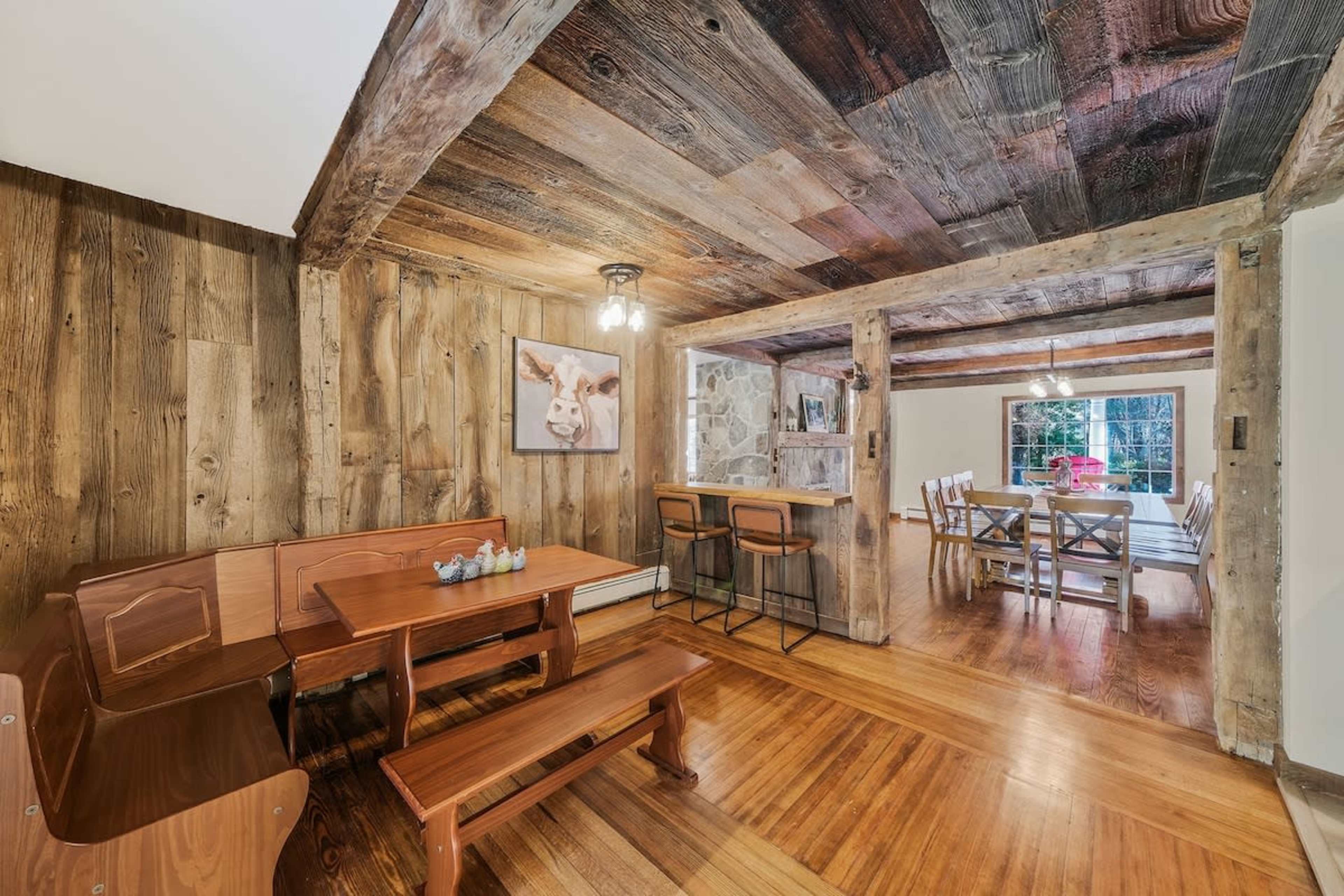 The image shows a rustic dining area with a wooden table and benches, featuring exposed wooden beams on the ceiling and a partially open layout leading to another dining space.