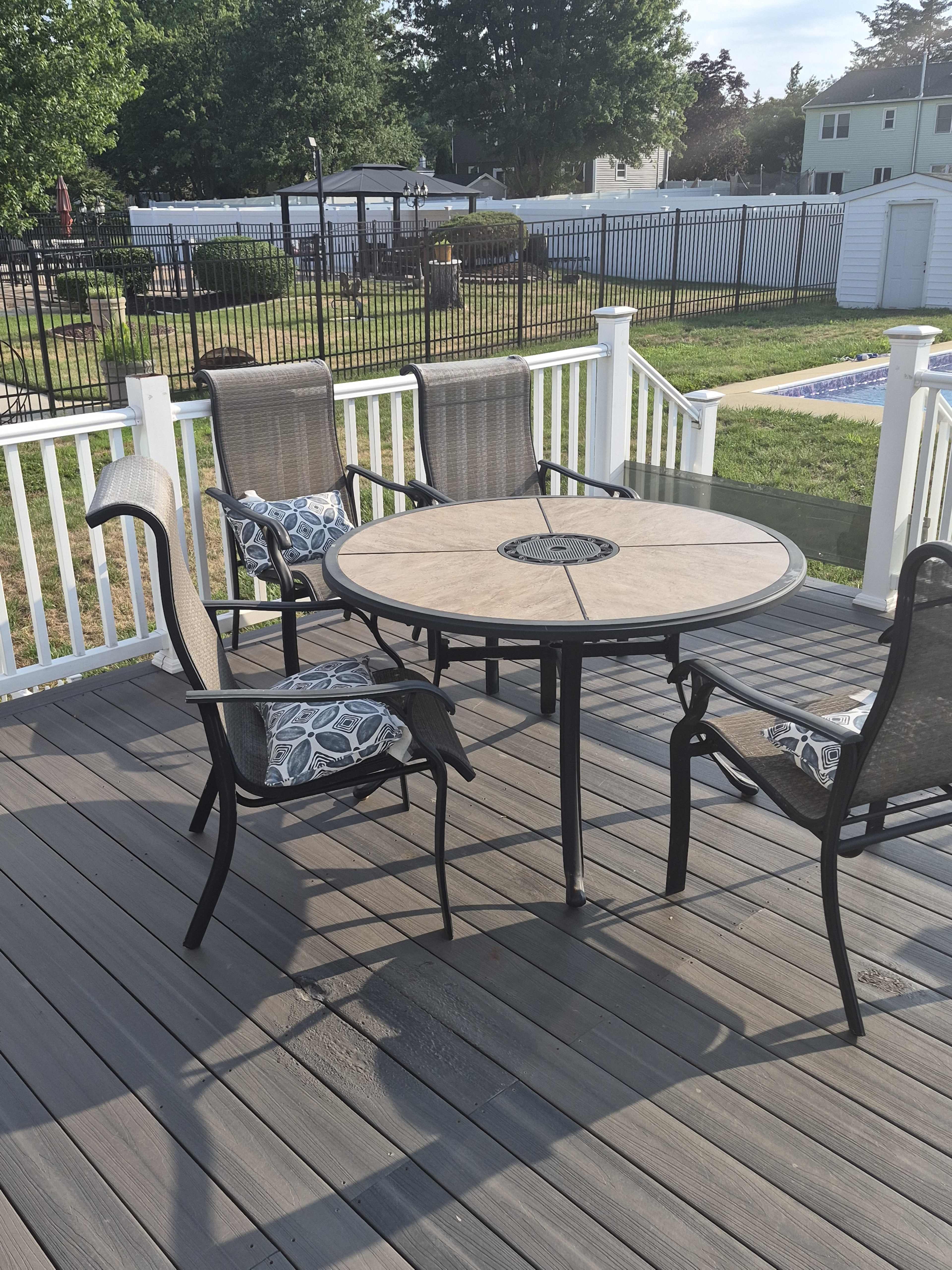 The image shows a patio set consisting of a round table and four chairs with cushions, situated on a wooden deck.