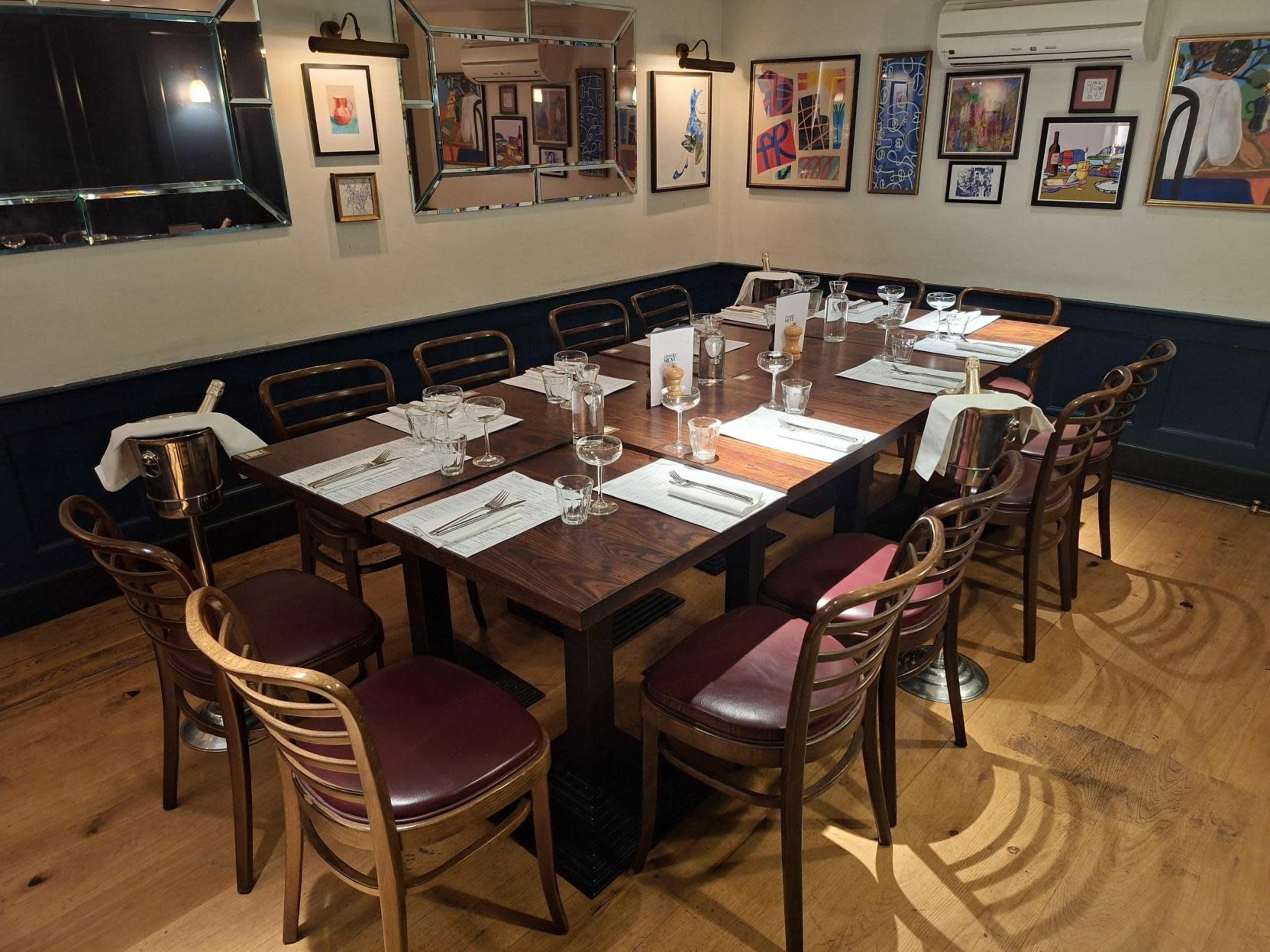 A long wooden table is set for dining in a restaurant, surrounded by chairs and decorated with framed artwork on the walls.