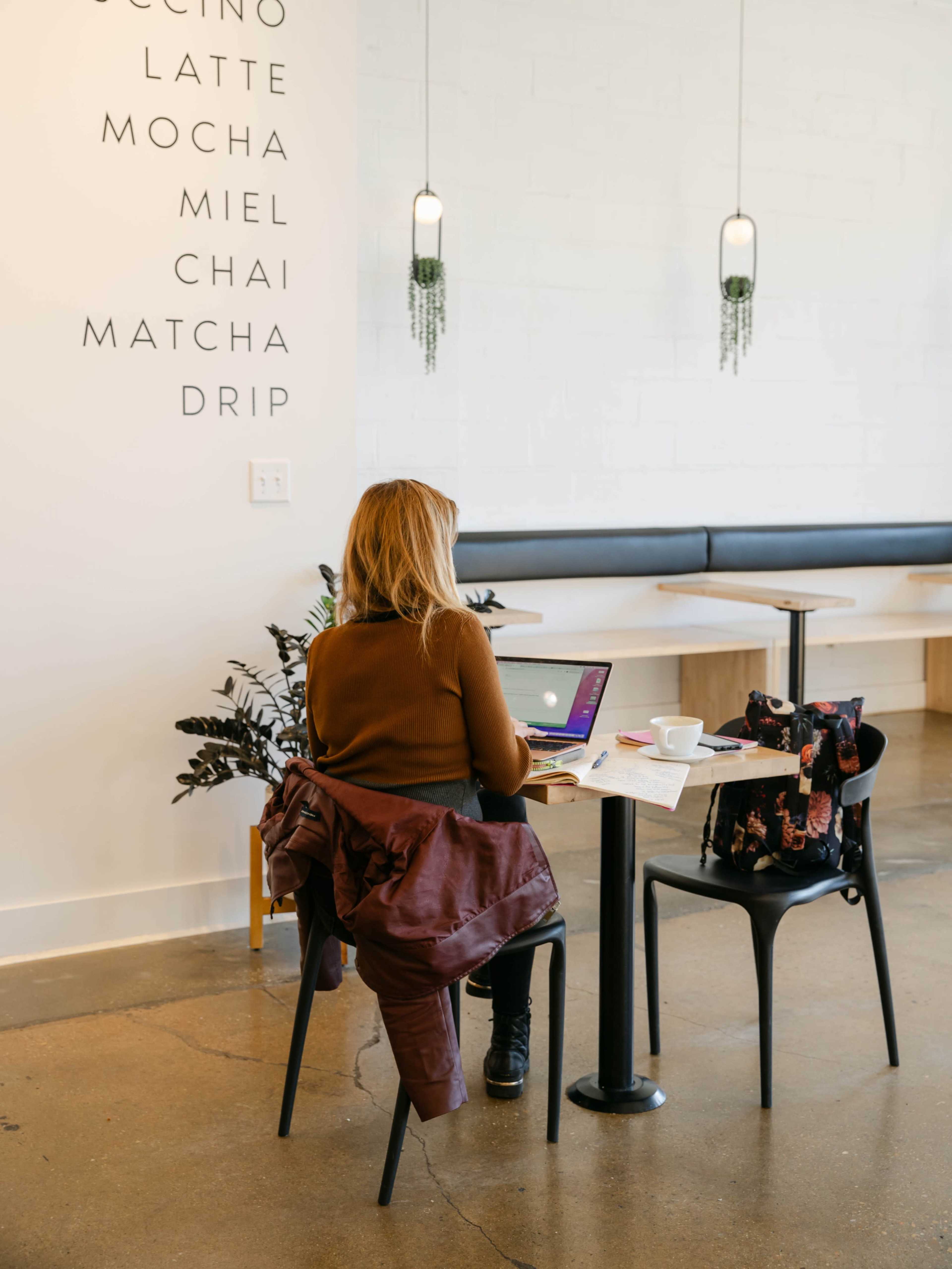 A woman sits at a small table in a café, working on a laptop with a notepad and drinks nearby.