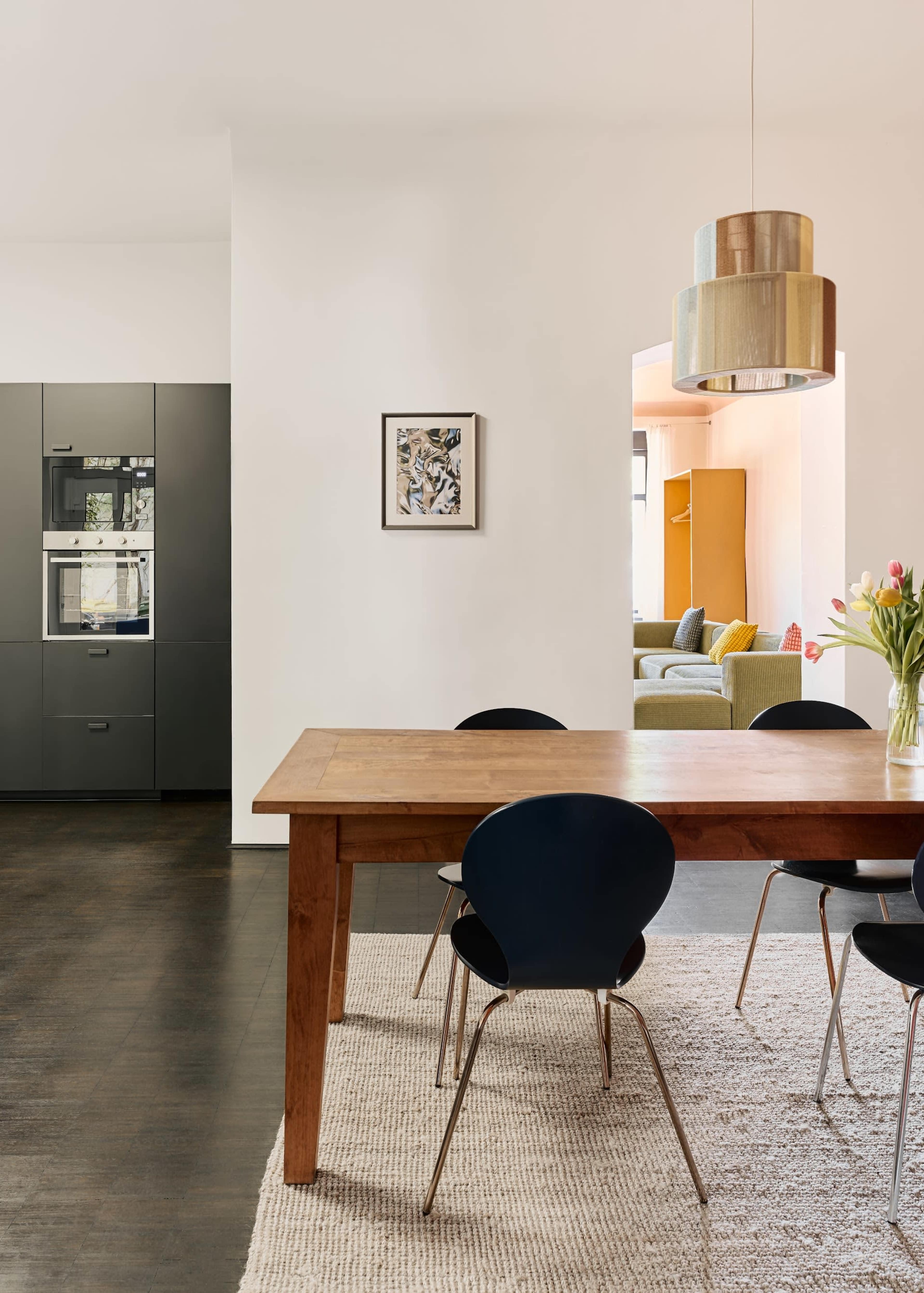 The image shows a modern kitchen and dining area featuring a wooden table with black chairs, a gray kitchen cabinet, and a light fixture hanging above the table.