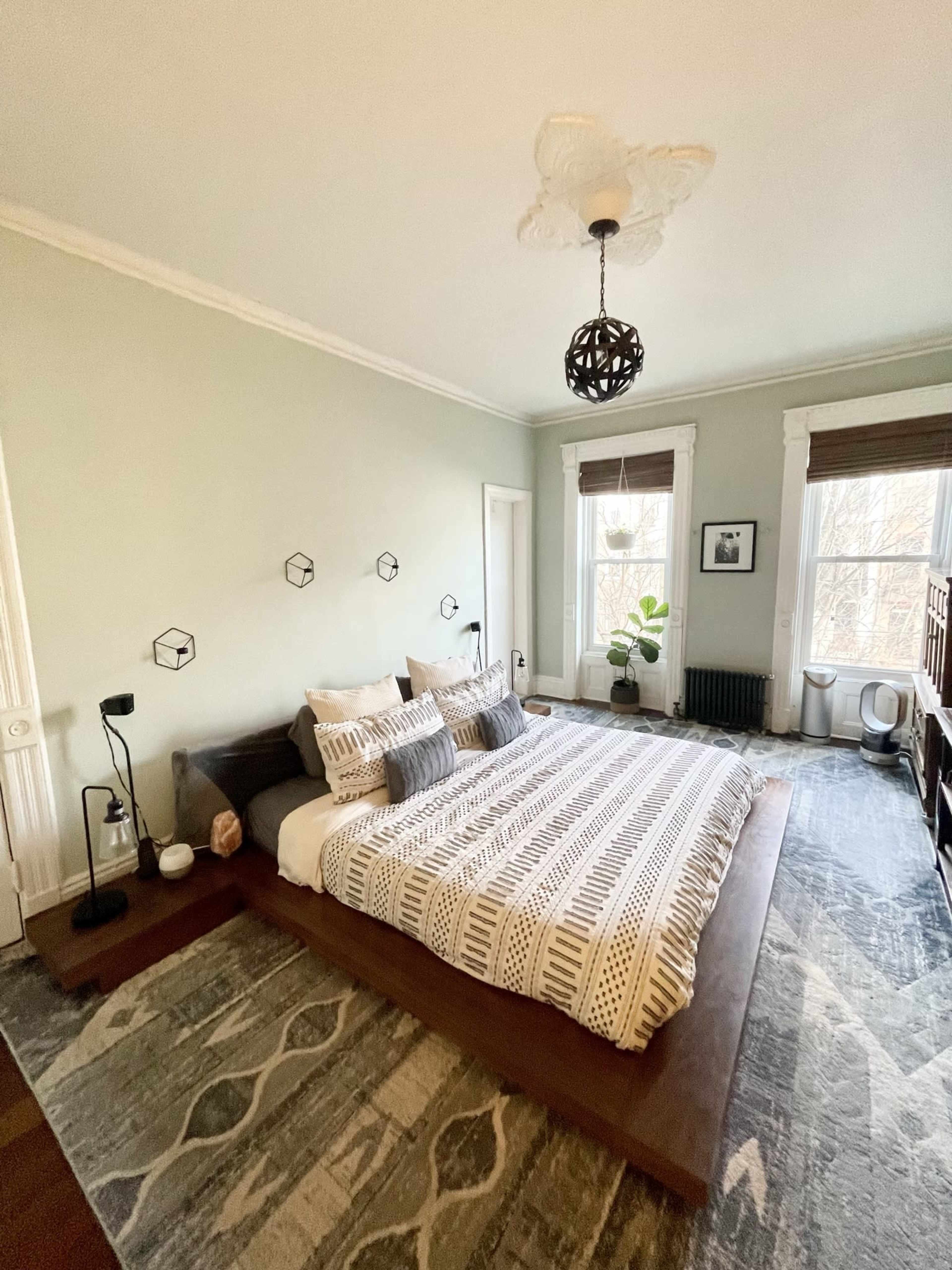 A spacious bedroom with a large bed, patterned bedding, and large windows allowing natural light, complemented by a green wall and wooden furniture.