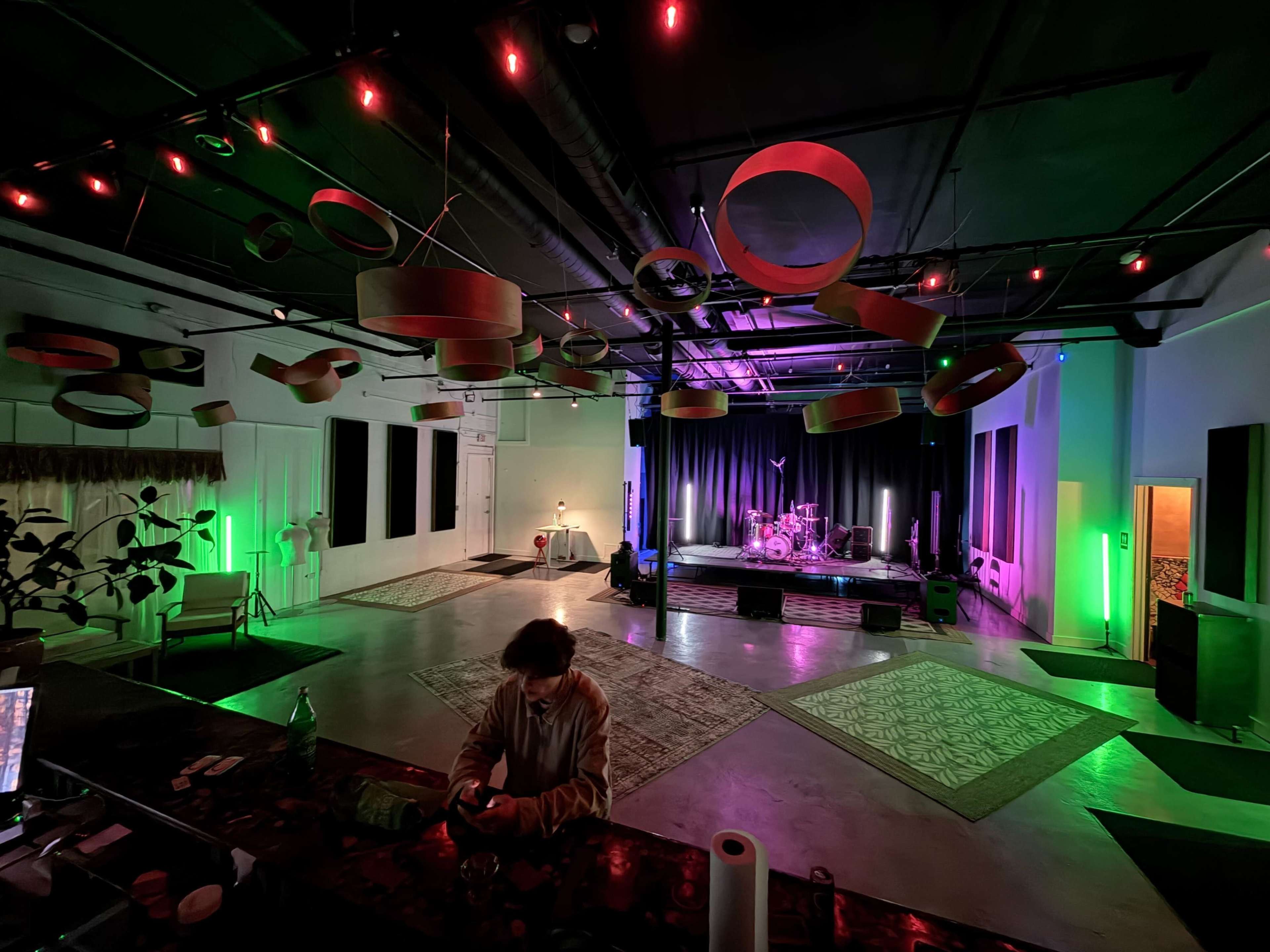 A spacious event venue features a stage with musical instruments, illuminated by colorful lights and adorned with hanging circular decorations.