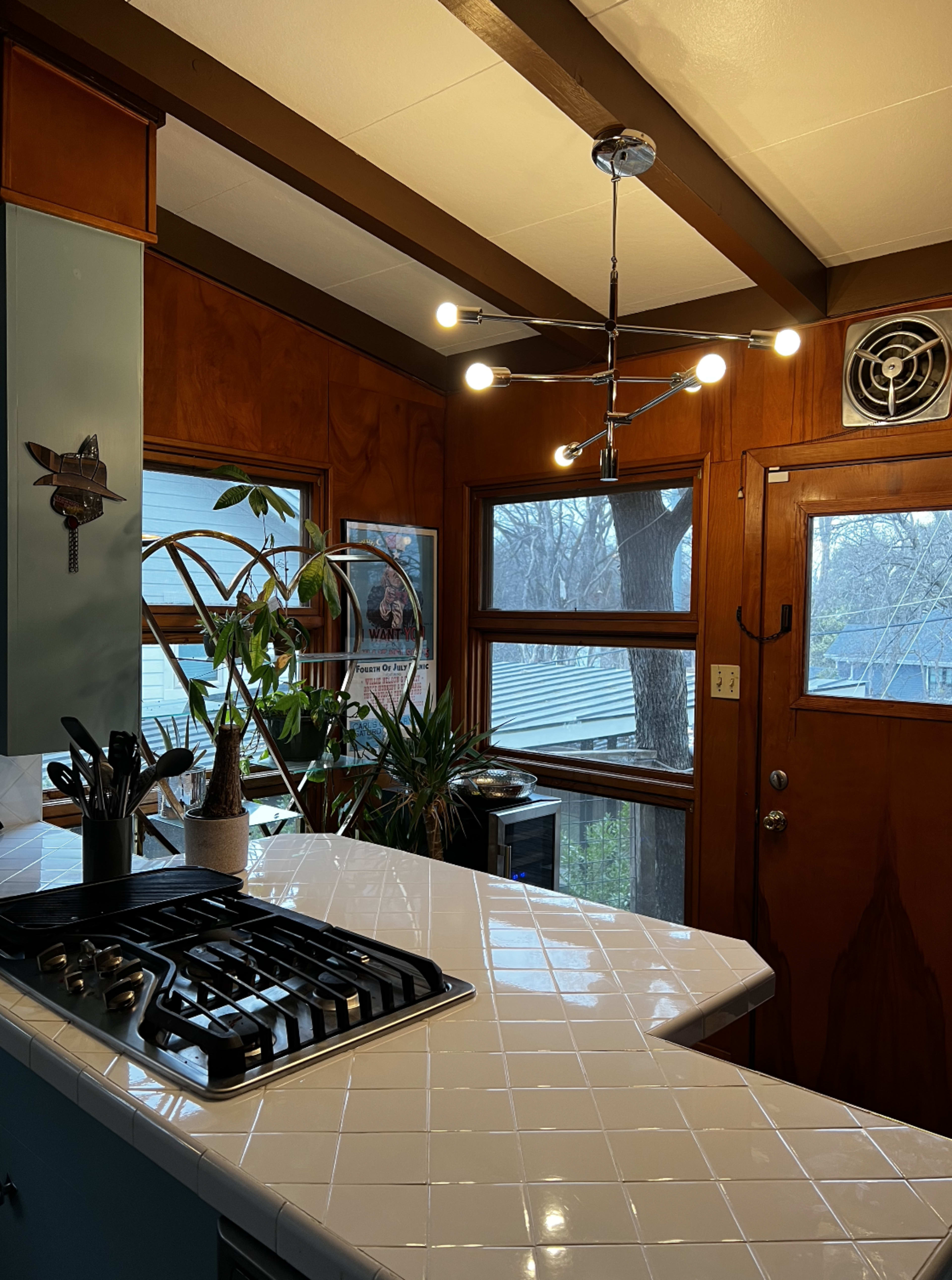 The image shows a kitchen with a tiled countertop, a modern light fixture, wooden walls, and large windows offering a view of trees outside.