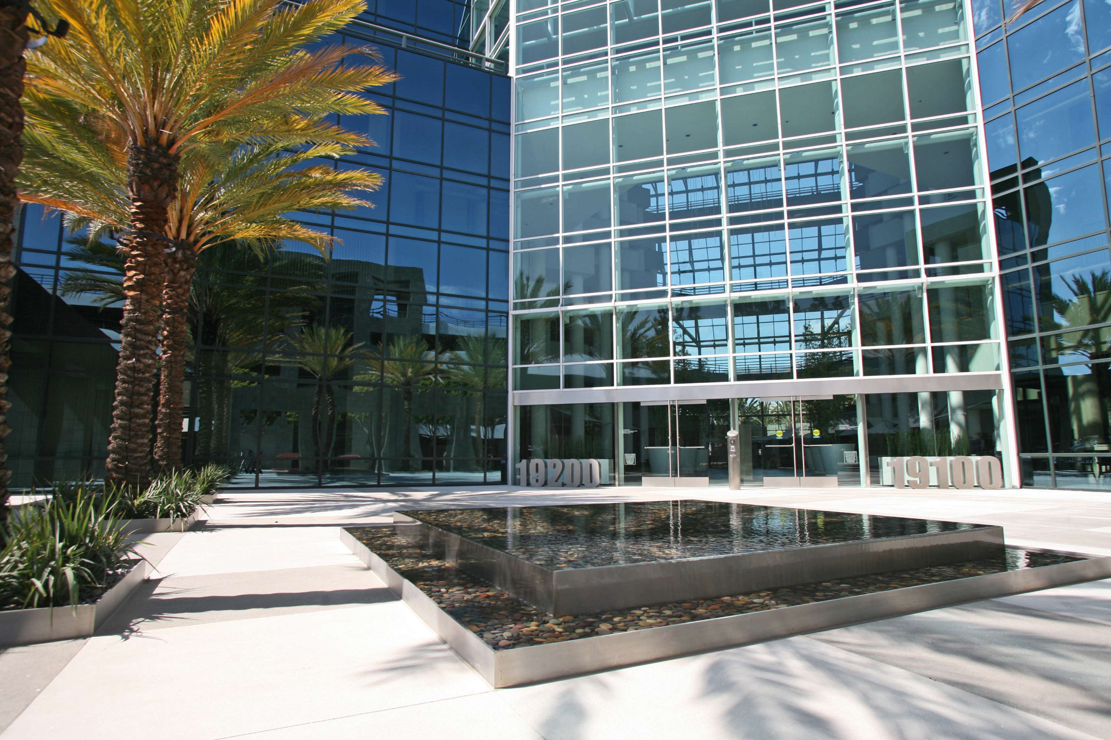 The image shows a modern glass building with palm trees and a reflective water feature in the courtyard.