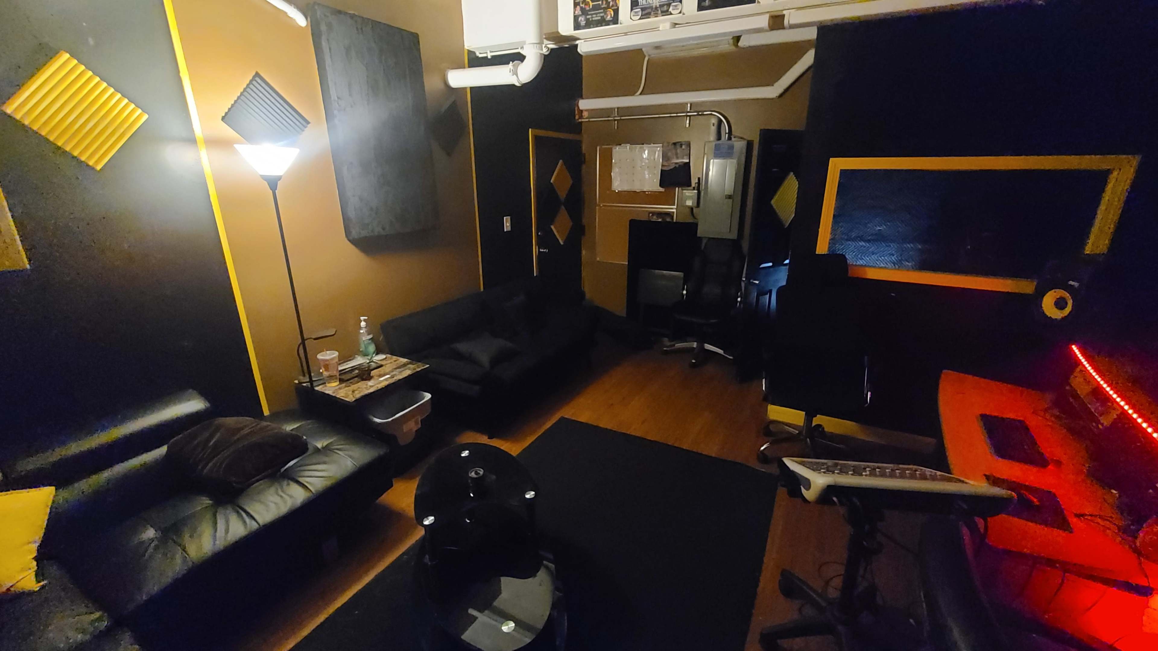 A small music studio with dark walls, couches, a lamp, a desk with equipment, and soundproofing panels on the walls.