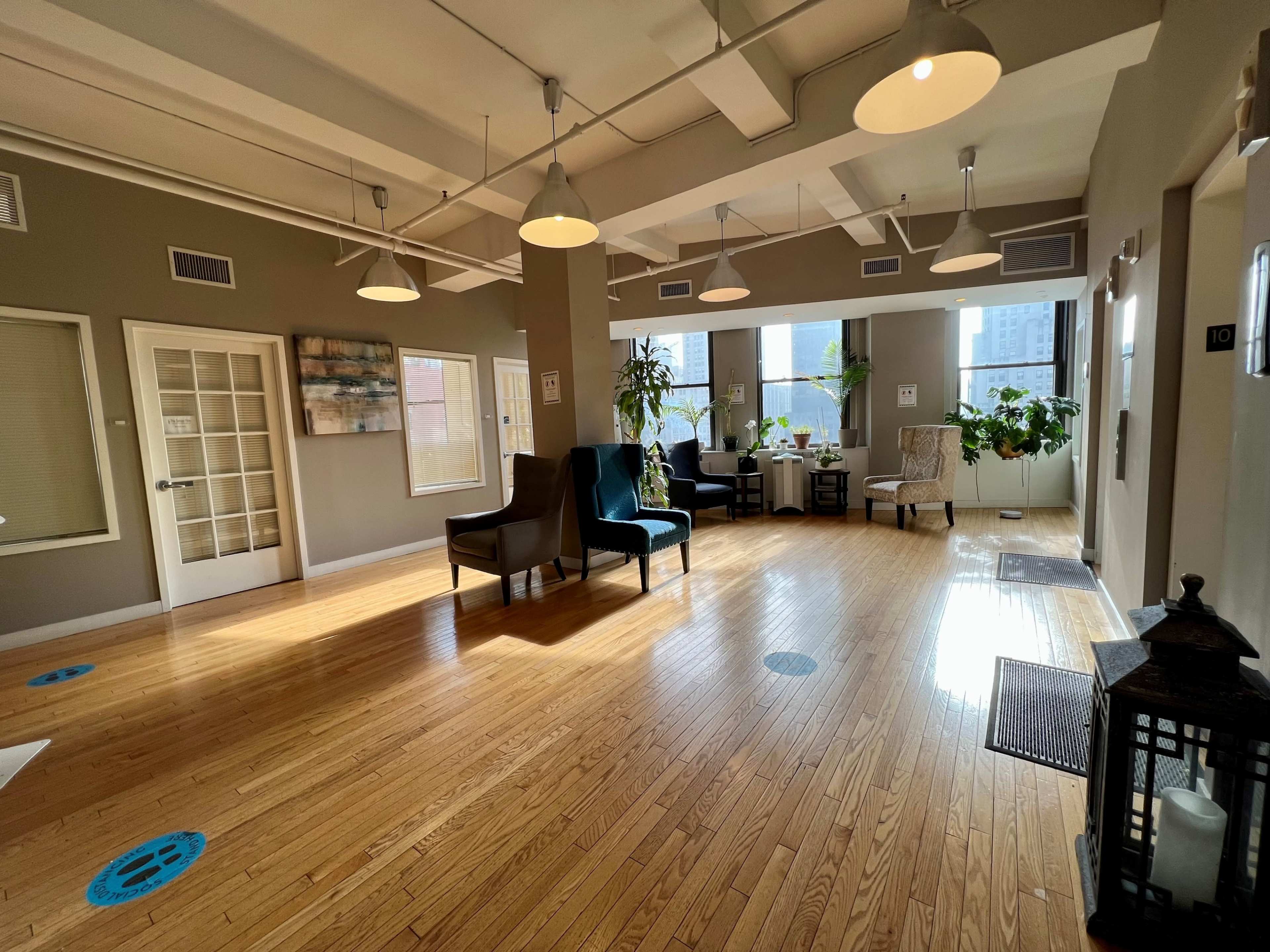 A spacious, well-lit common area with wooden flooring, seating arrangements, plants, and a reception desk.