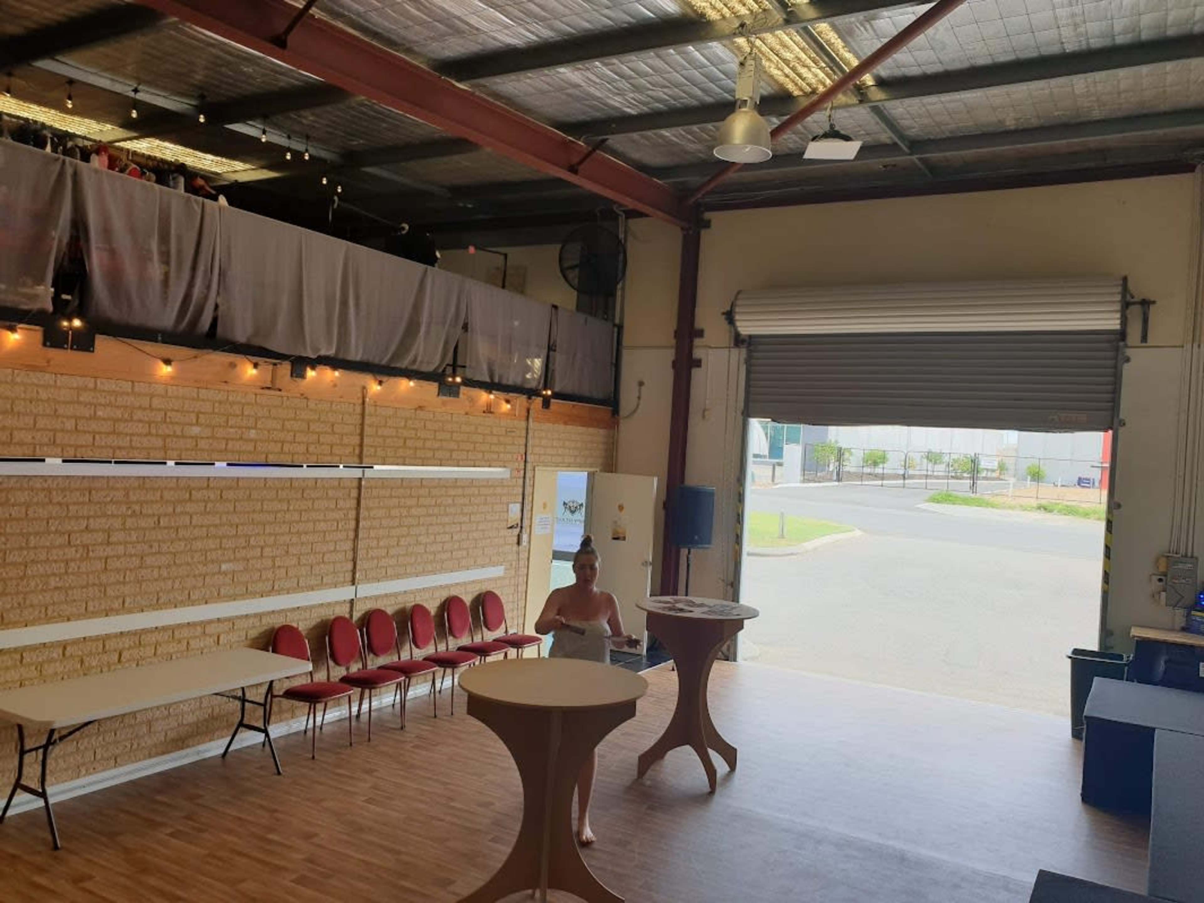 An indoor event space with a woman standing near round tables, overlooking rows of red chairs and a large open door leading outside.