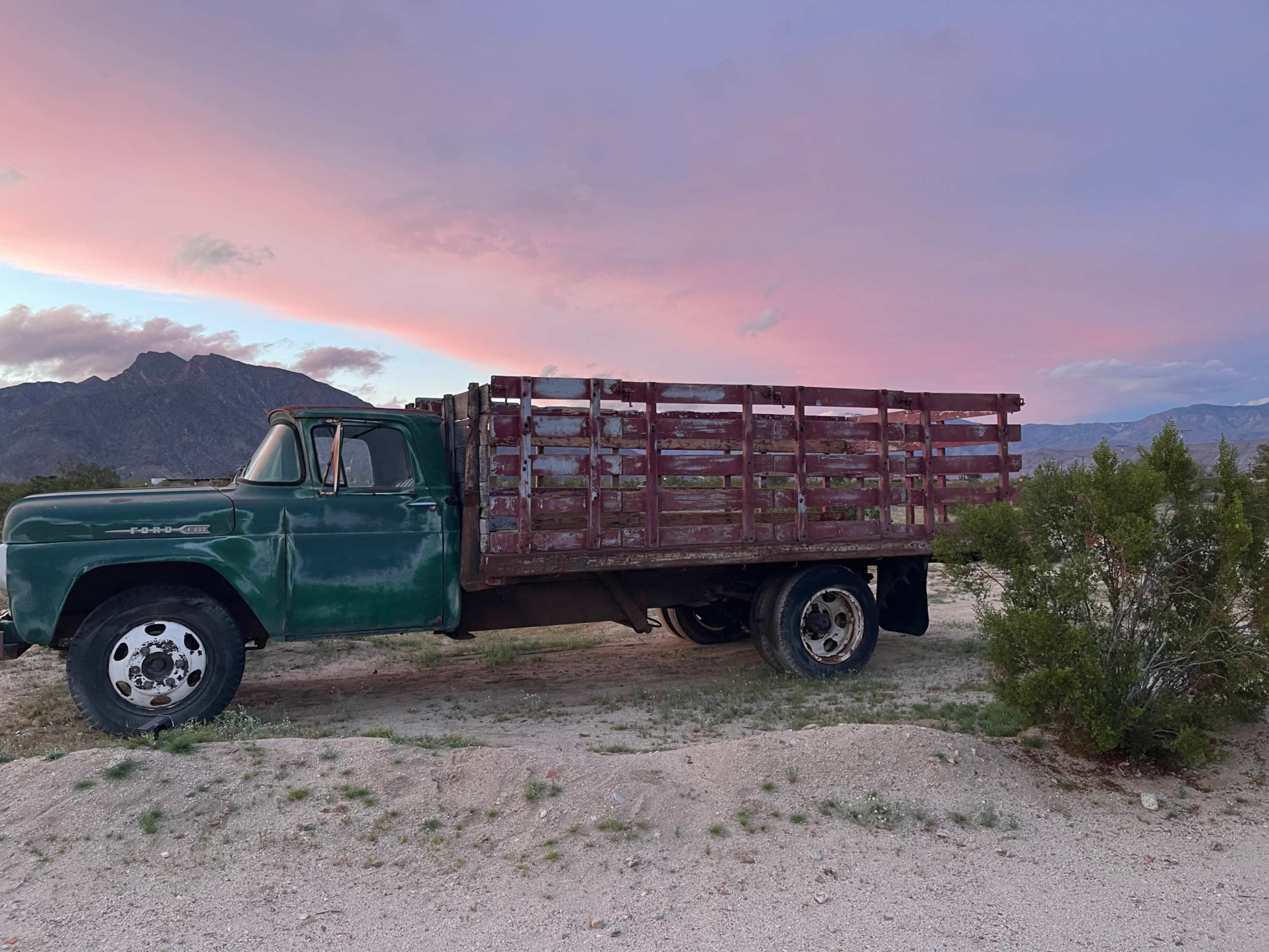 An old green truck with a wooden flatbed is parked on a gravel surface with mountains and a colorful sky in the background.