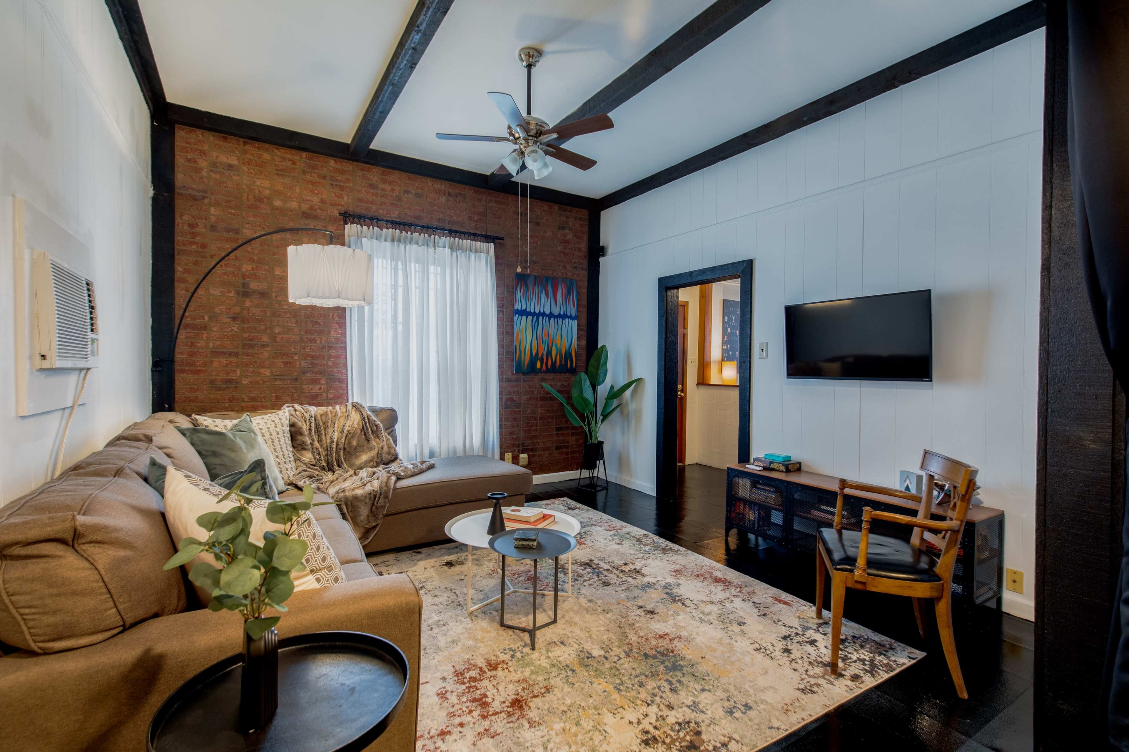 The image shows a cozy living room with a sectional sofa, a circular coffee table, a flat-screen TV, and a plant, all set against a backdrop of brick and wooden beams.