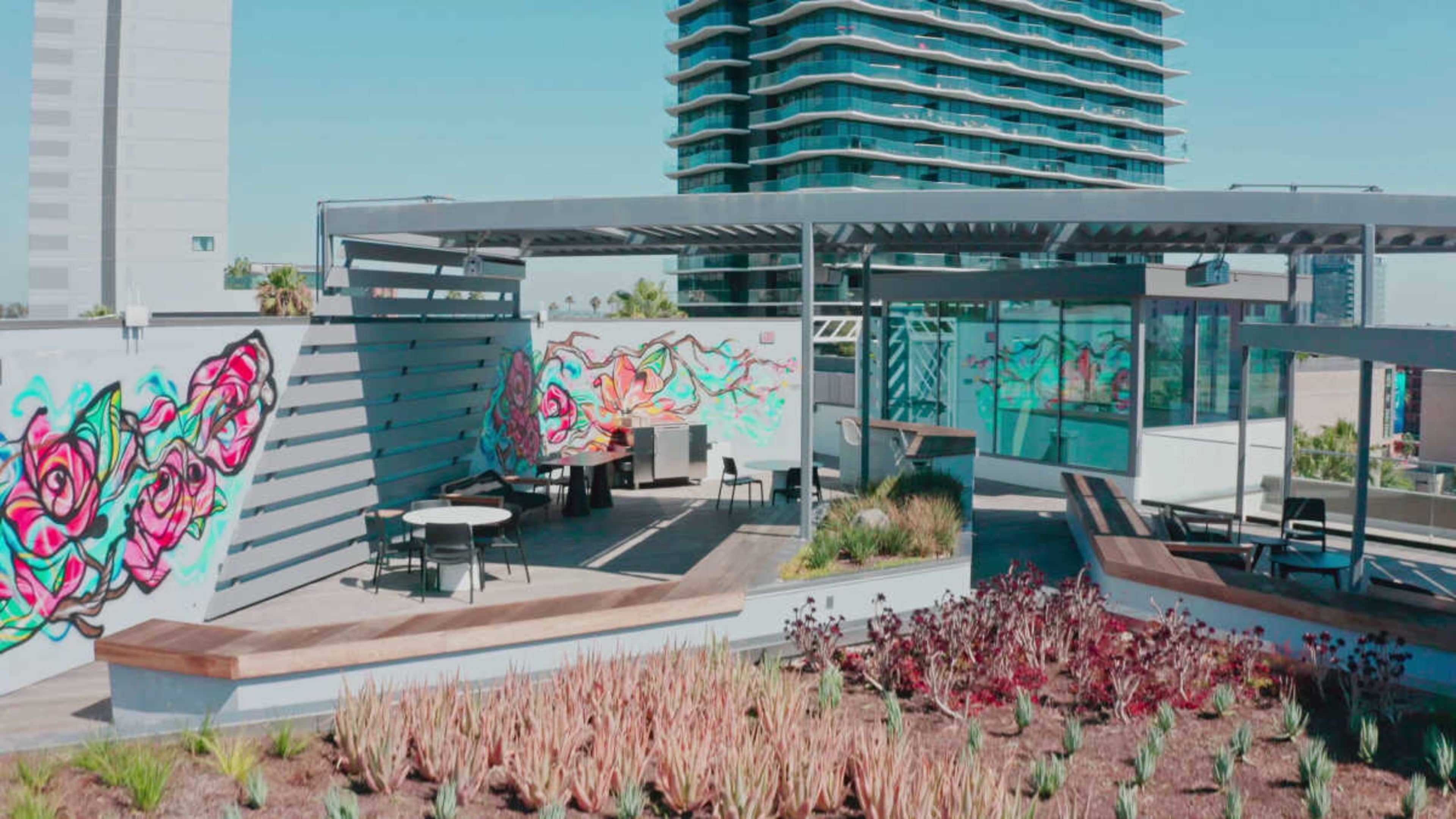 The image shows a rooftop space featuring vibrant mural art, modern seating, and landscaped greenery, with a view of a multistory building in the background.