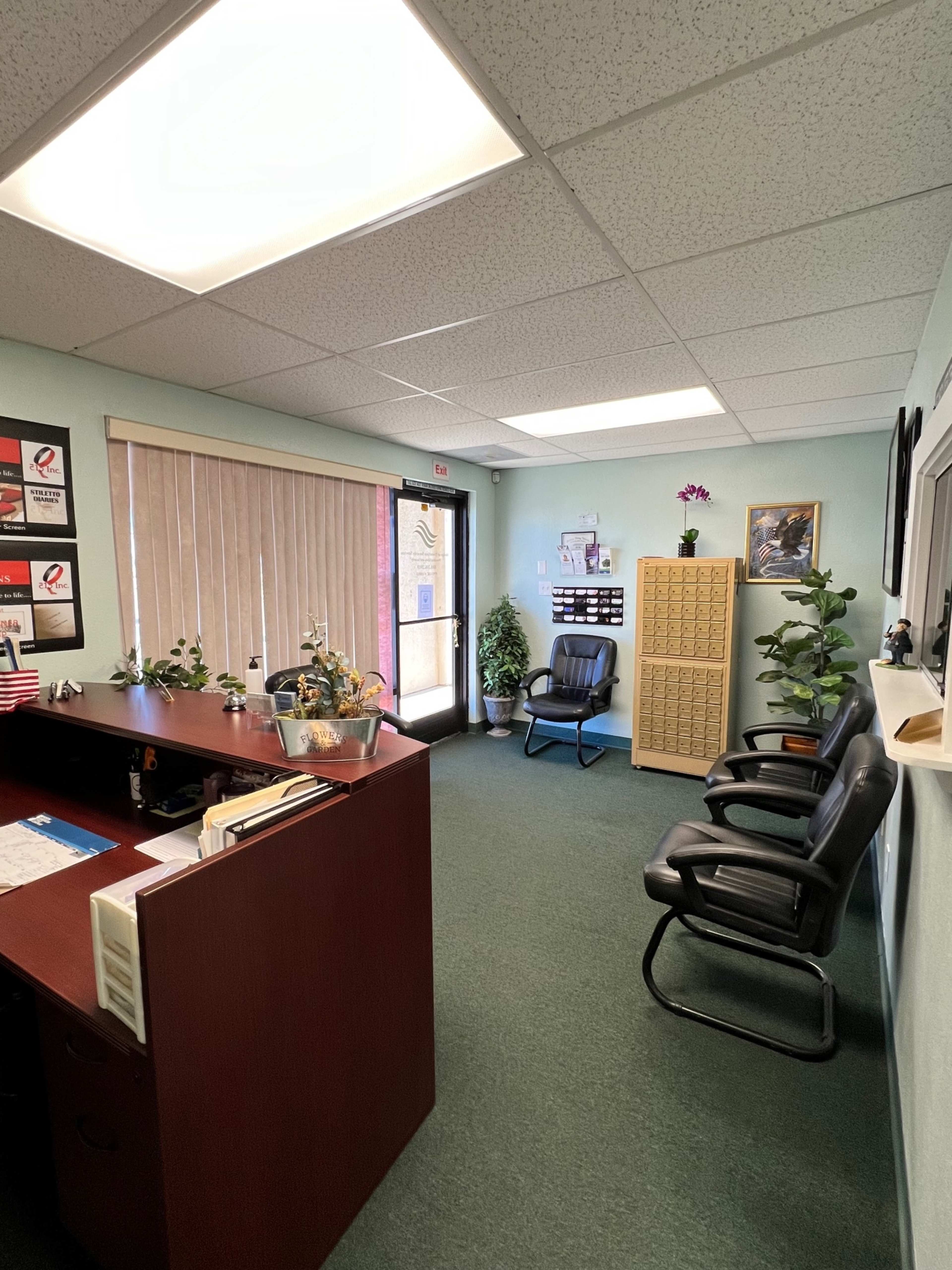 A waiting area in an office with a reception desk, chairs, indoor plants, and a window with blinds.