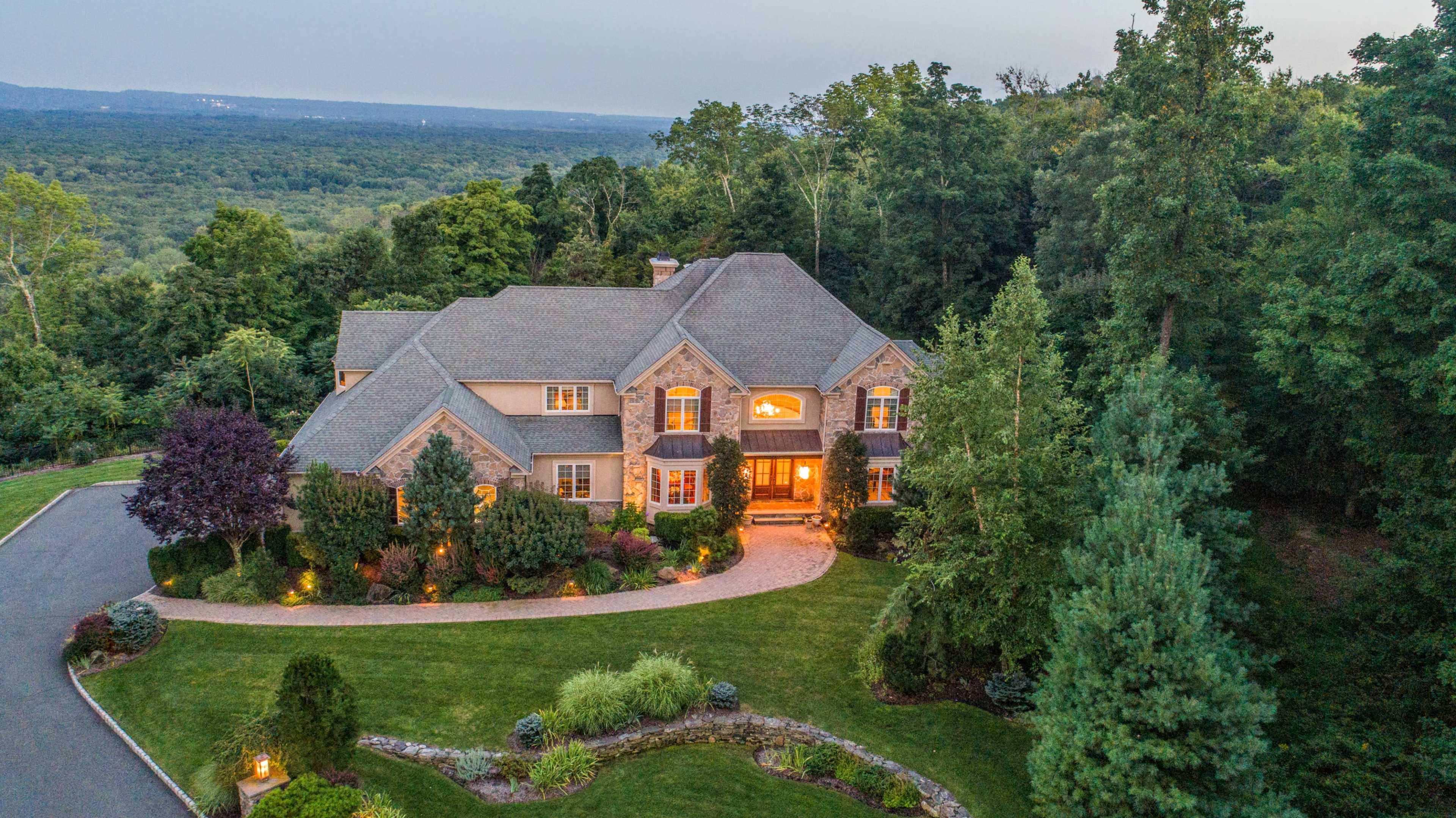 A large, multi-story stone and shingle house is set on a landscaped property surrounded by trees and hills, with warm lights illuminating the entrance.