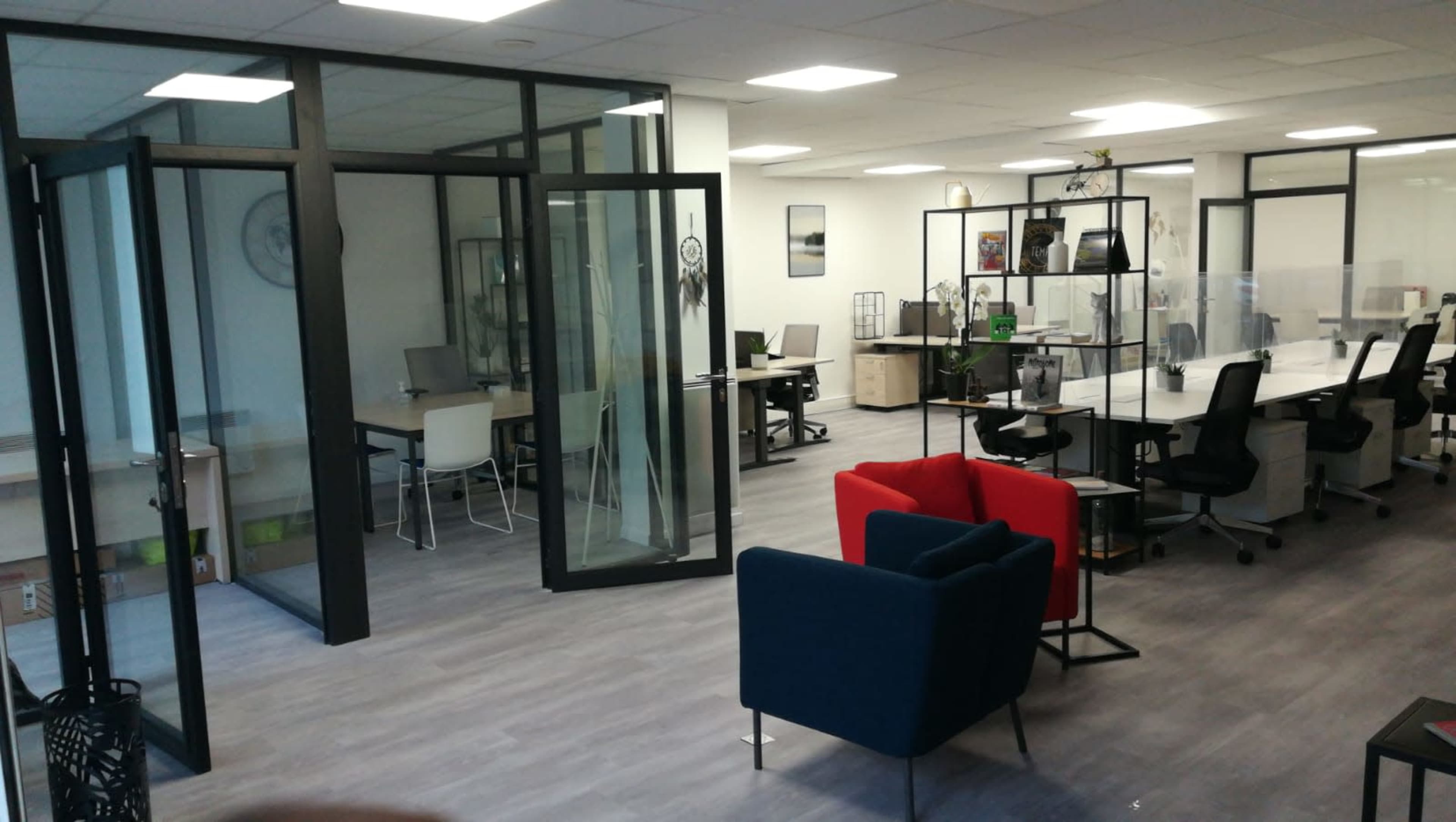 A modern office space featuring glass partitioned areas, several desks with chairs, and a seating area with red and blue couches.