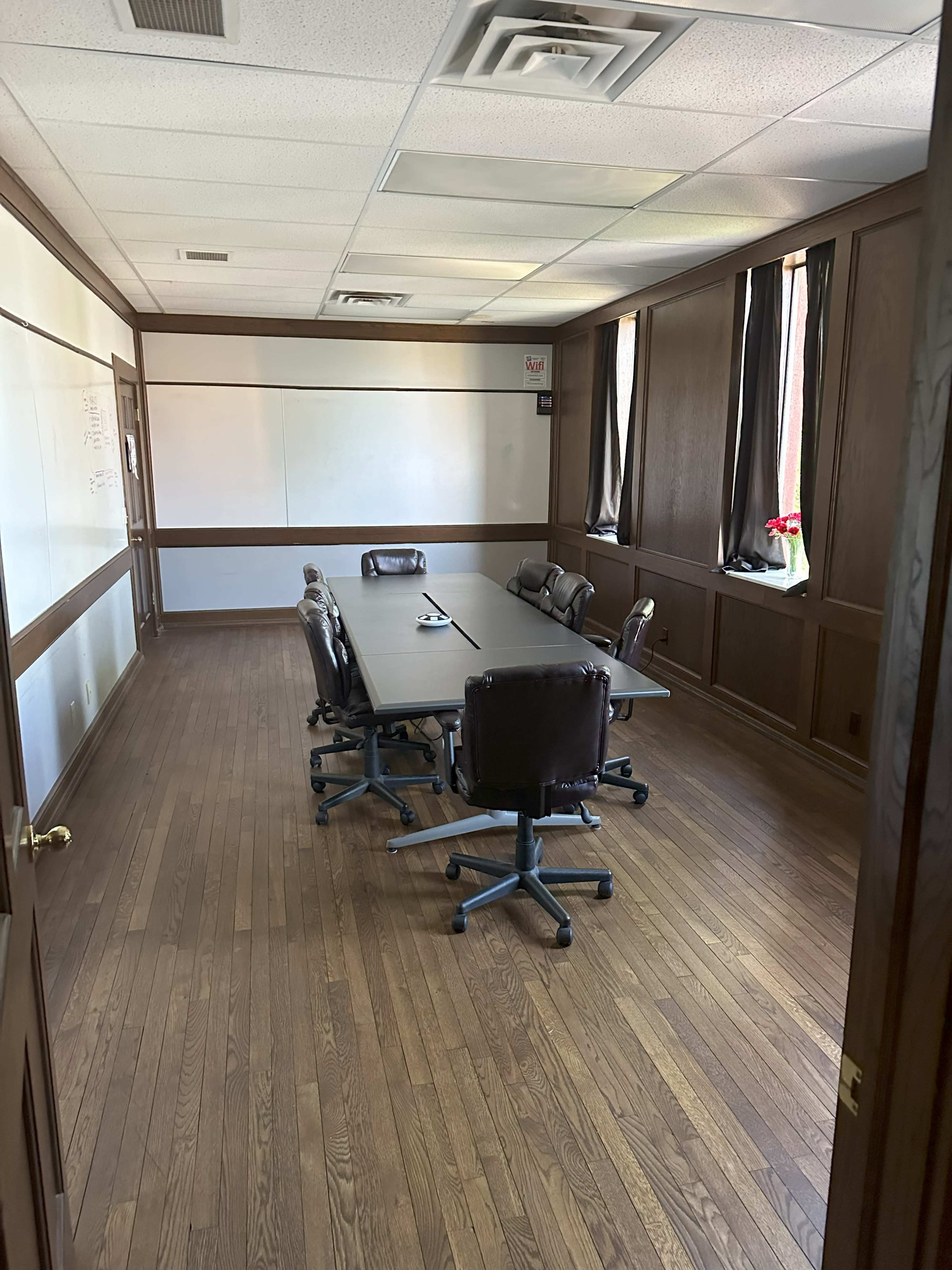 A conference room with a long table and several rolling chairs, featuring wooden paneling and large windows.