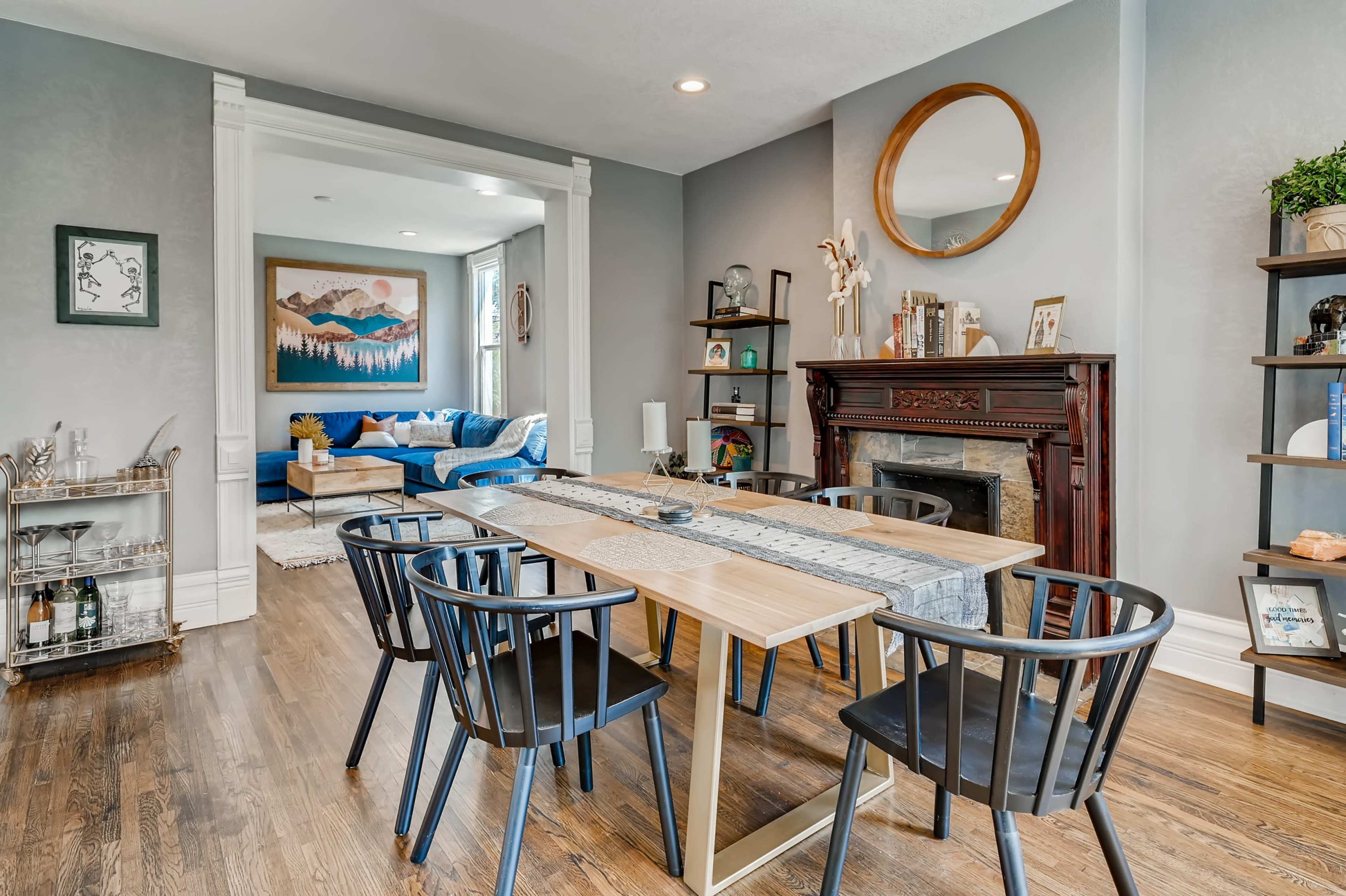 A dining area features a wooden table with black chairs, a decorative mirror on the wall, and a cozy living room visible in the background.