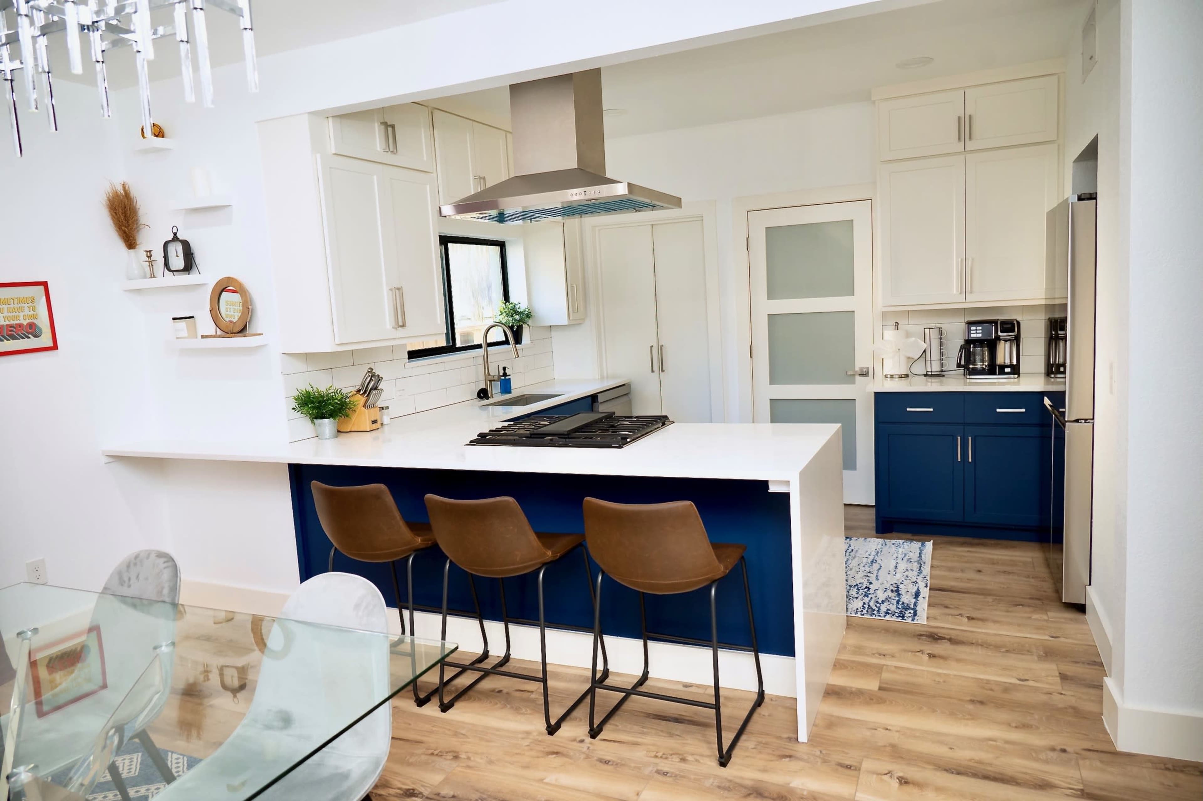 The image shows a modern kitchen featuring a white and navy blue color scheme, with a kitchen island that includes bar stools and a stainless steel hood above the stovetop.