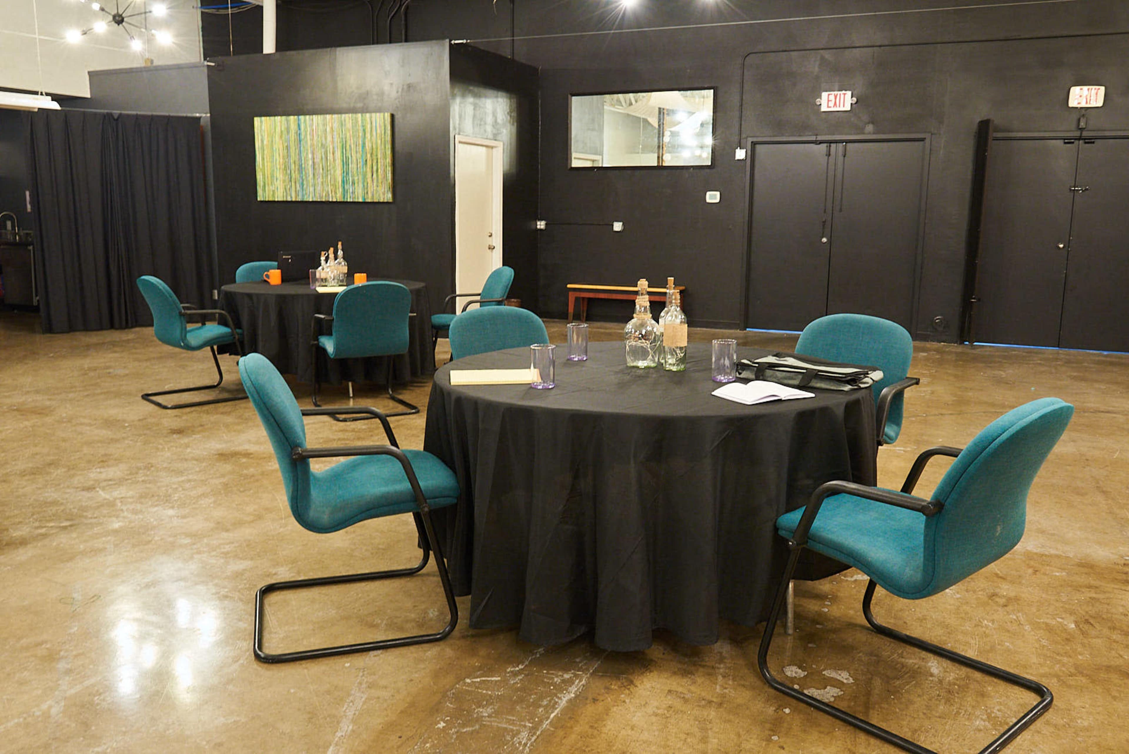 A room with several round tables covered in black tablecloths, each with turquoise chairs, a few bottles, and glasses, set against dark walls.