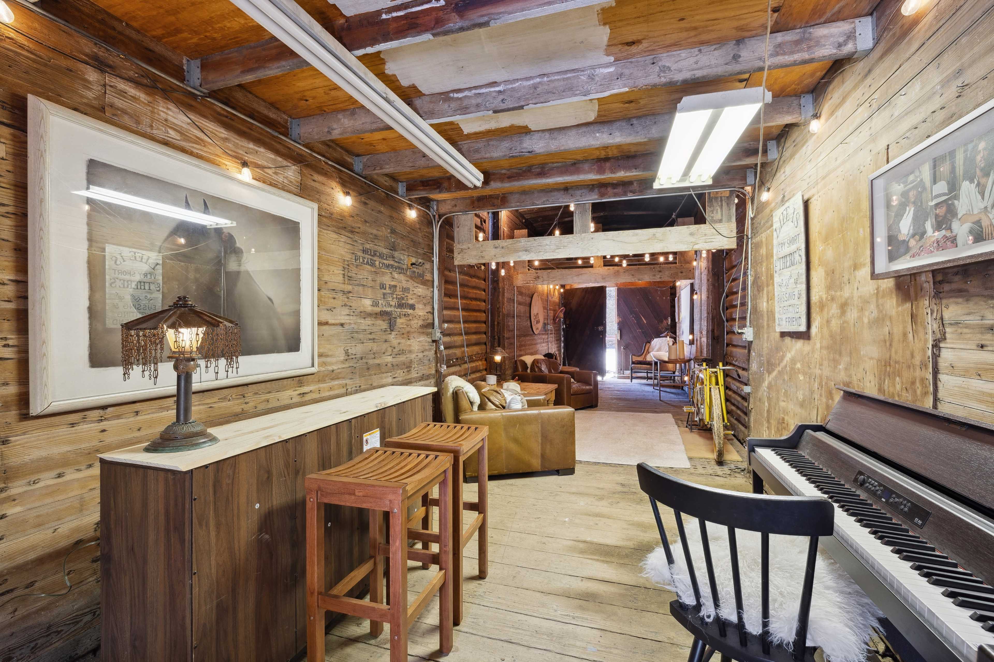 The image shows a rustic interior of a wooden structure with exposed beams, featuring a bar area with stools, a piano, and a comfortable seating area in the background.