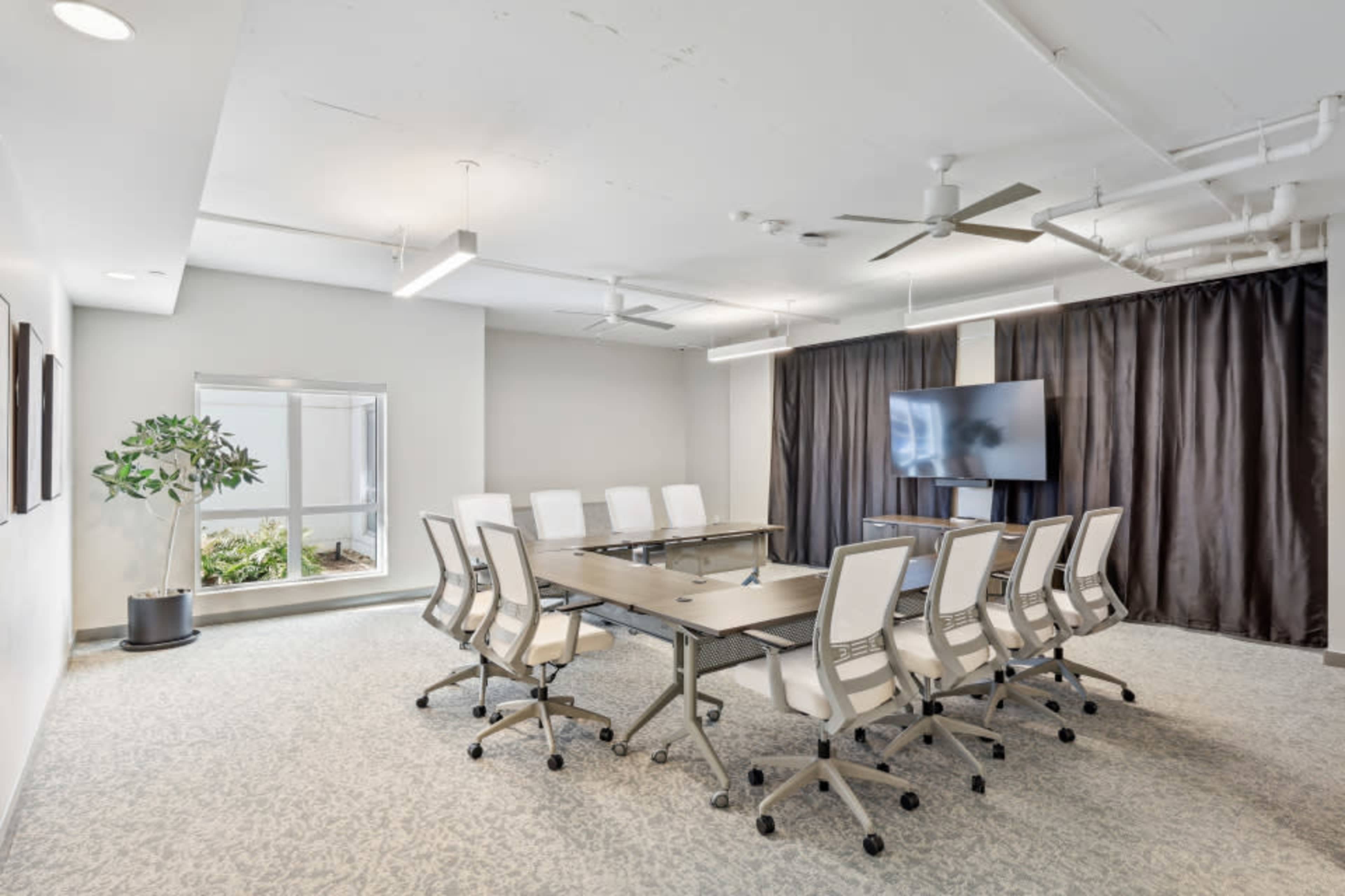 A modern conference room features a long table surrounded by white chairs, with a large screen mounted on a wall and a large window providing natural light.