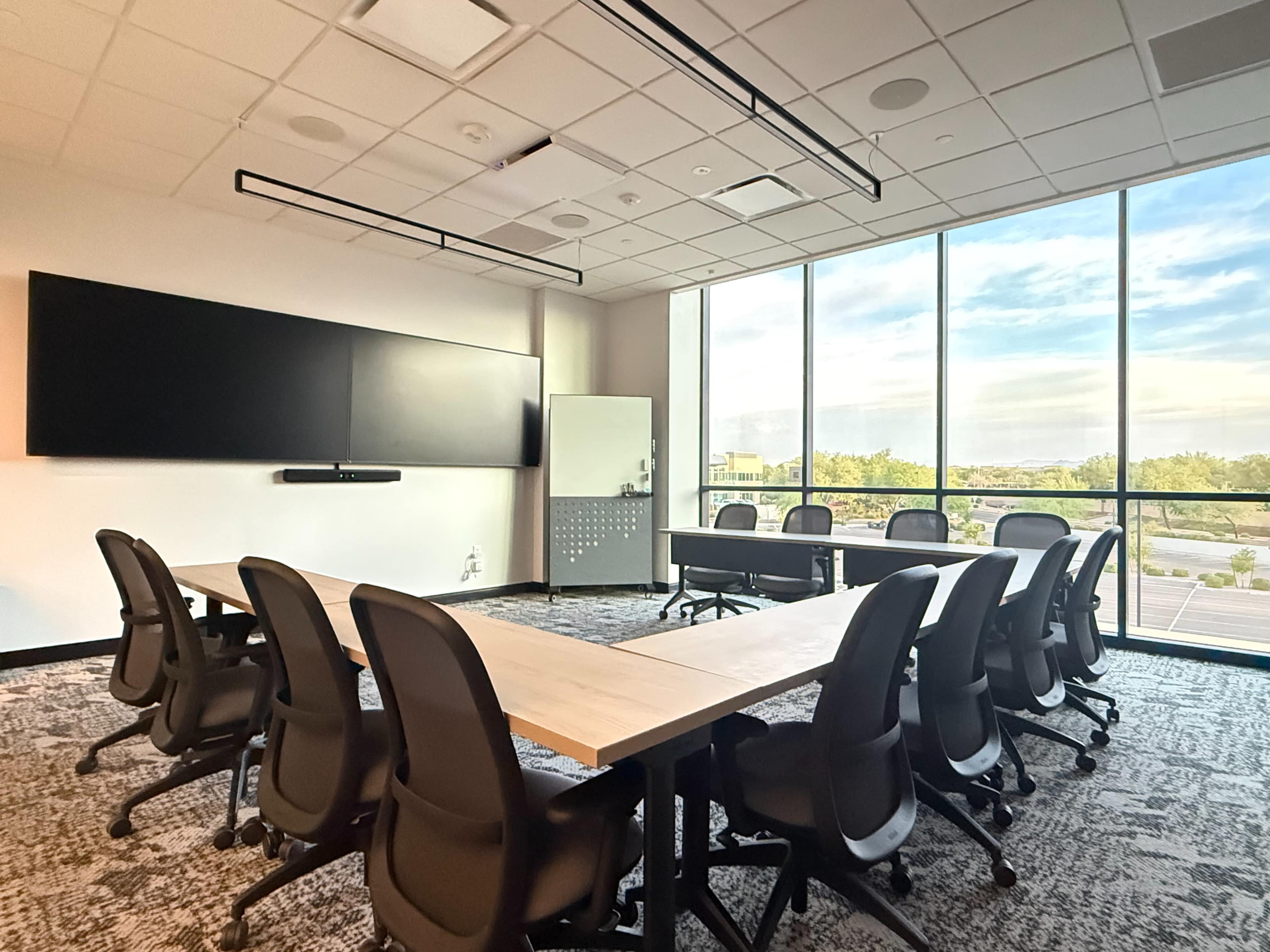 The image depicts a modern conference room with a large table, several chairs, and floor-to-ceiling windows offering a view of the outside.