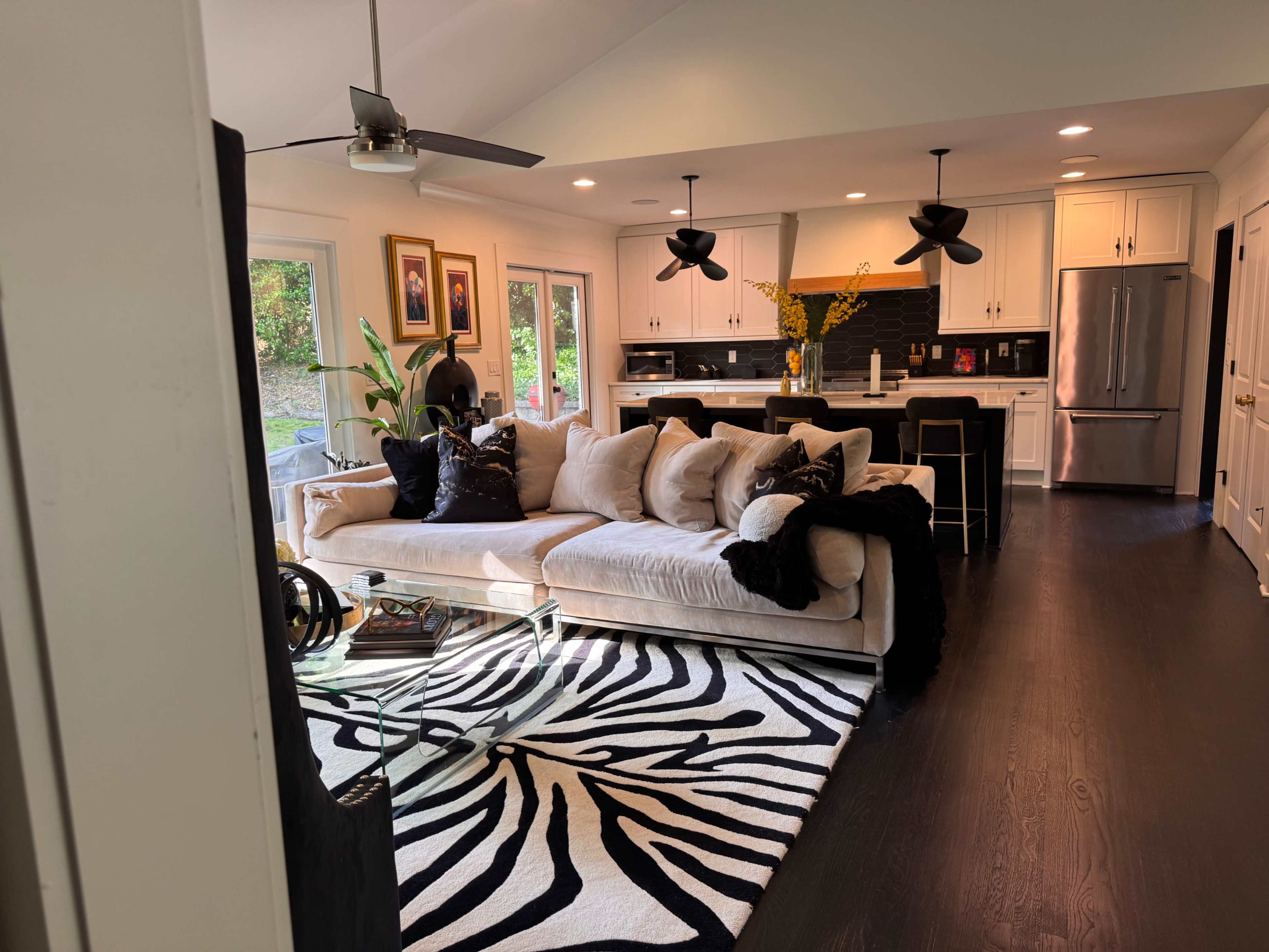 The image shows a modern living room featuring a large beige sectional sofa on a black and white striped area rug, with a glass coffee table and a kitchen visible in the background.