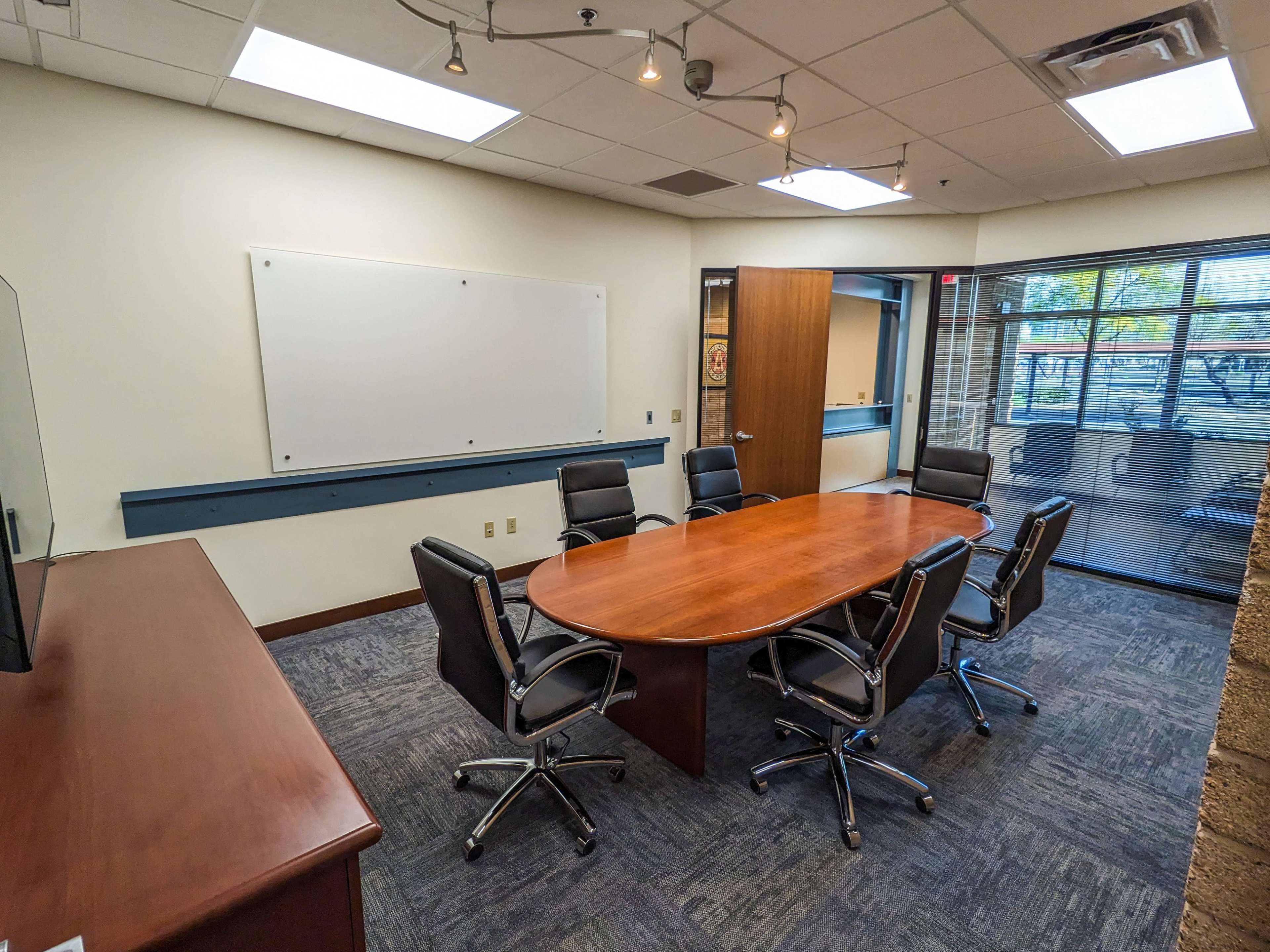 A conference room features a large wooden table surrounded by six black chairs, with a whiteboard and a TV screen on the wall.