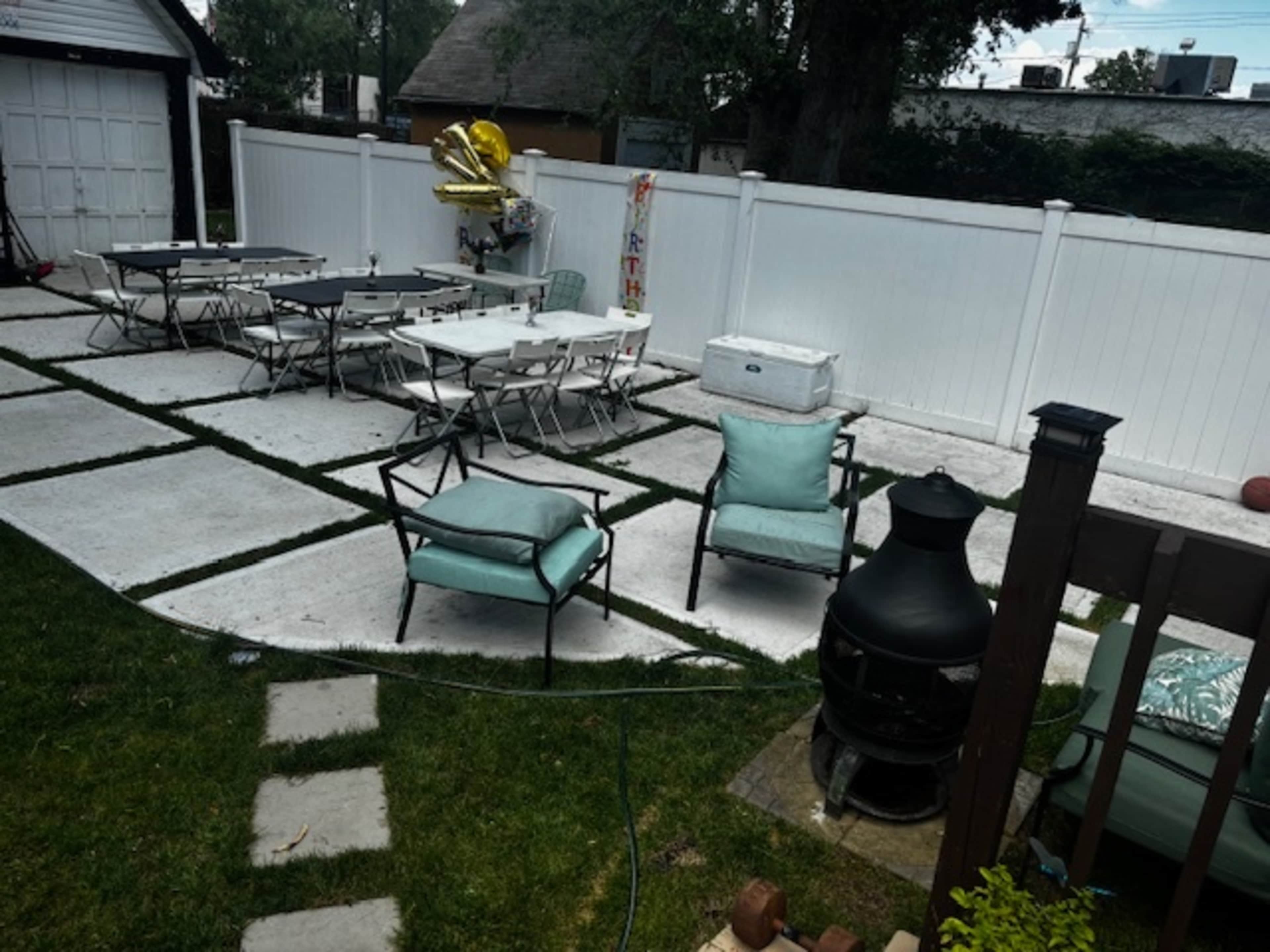 A backyard patio area with a mix of concrete and grassy sections, featuring tables and chairs arranged for an event, a cooler, and a decorative balloon.