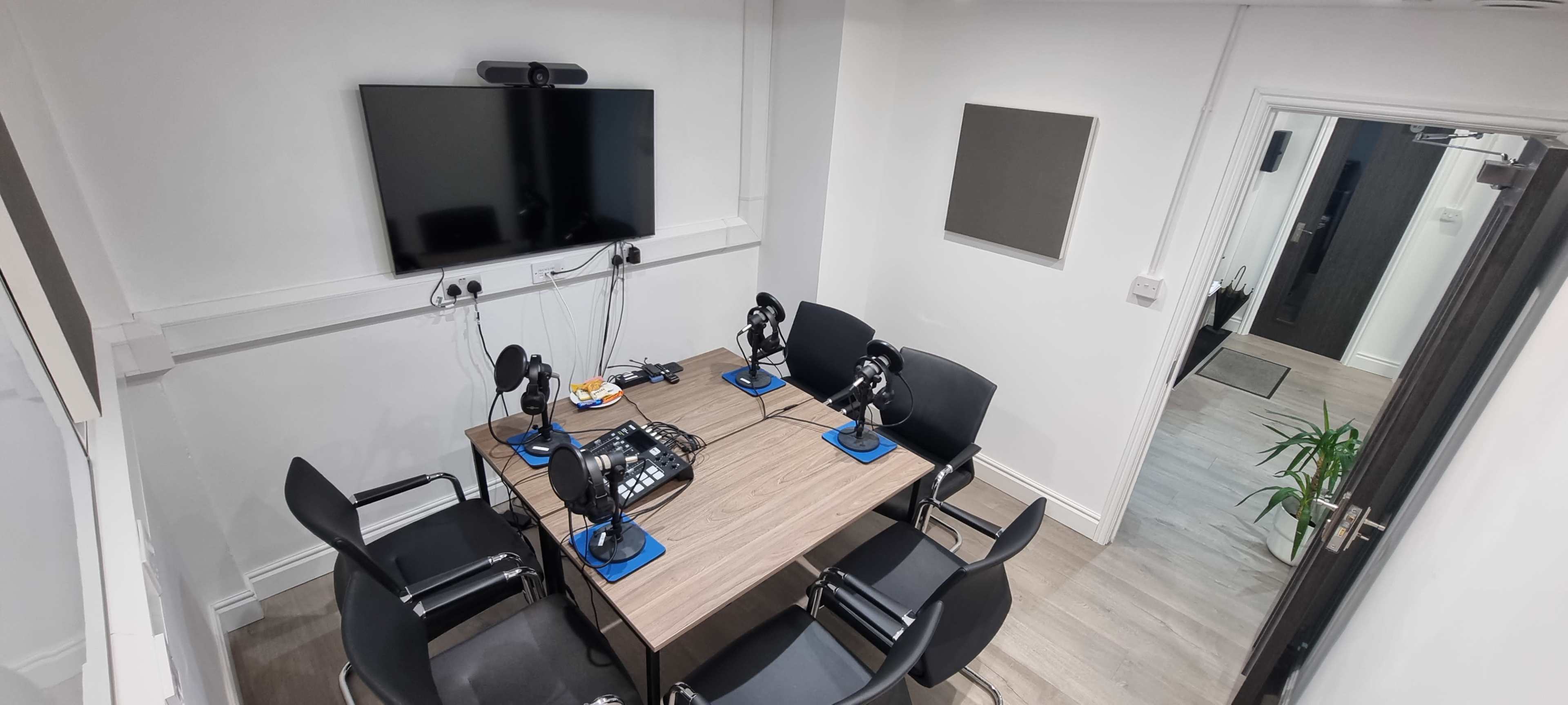 A soundproof meeting room features a wooden table with microphones and chairs arranged around it, along with a large television mounted on the wall.