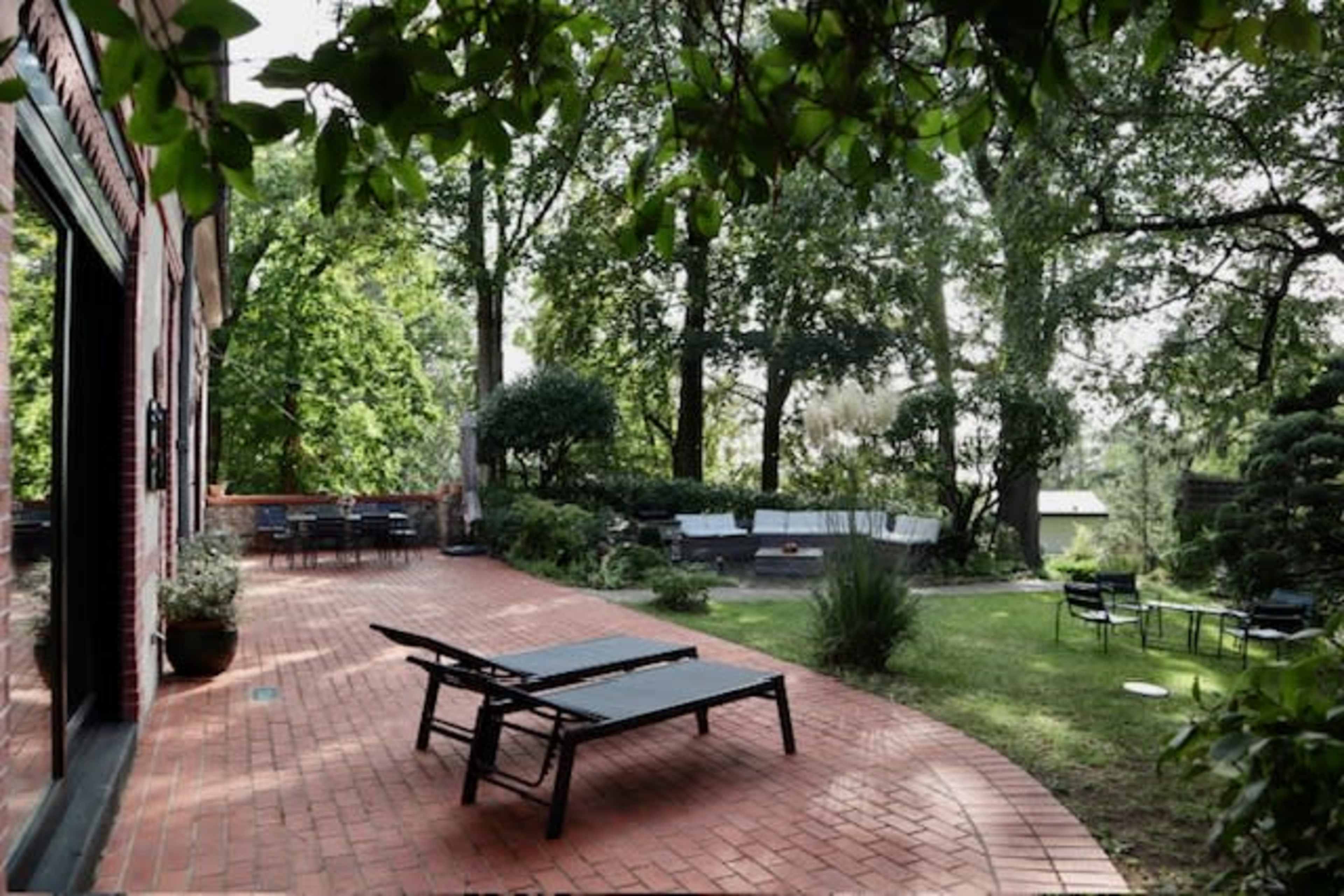 A landscaped backyard with brick pathways, lounge chairs, and a seating area surrounded by trees.