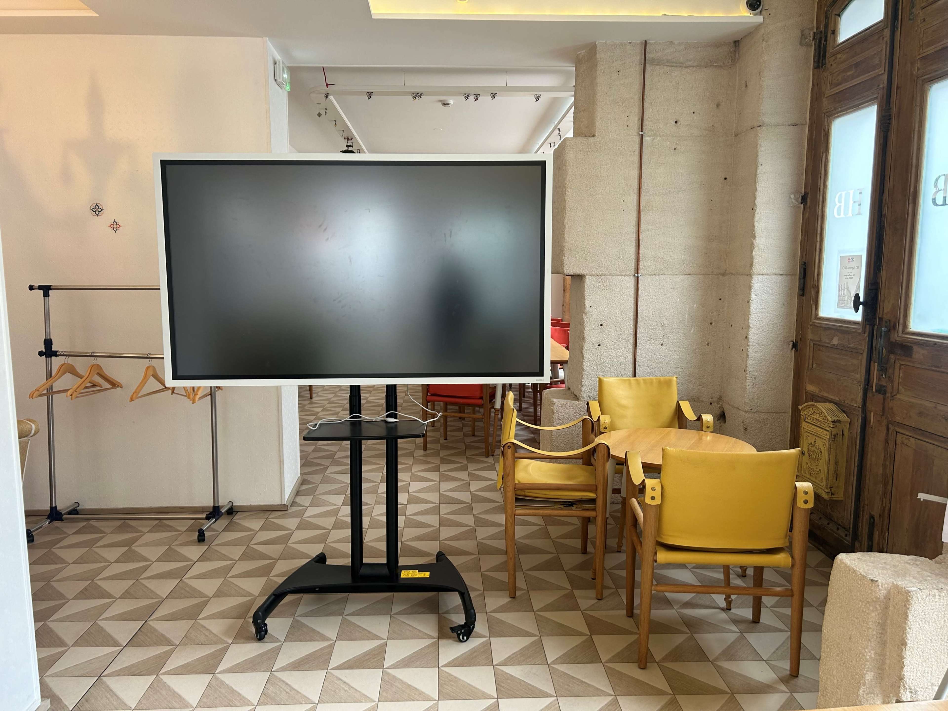 The image shows a large digital display on a wheeled stand next to a small round table and yellow chairs in a room with patterned flooring.