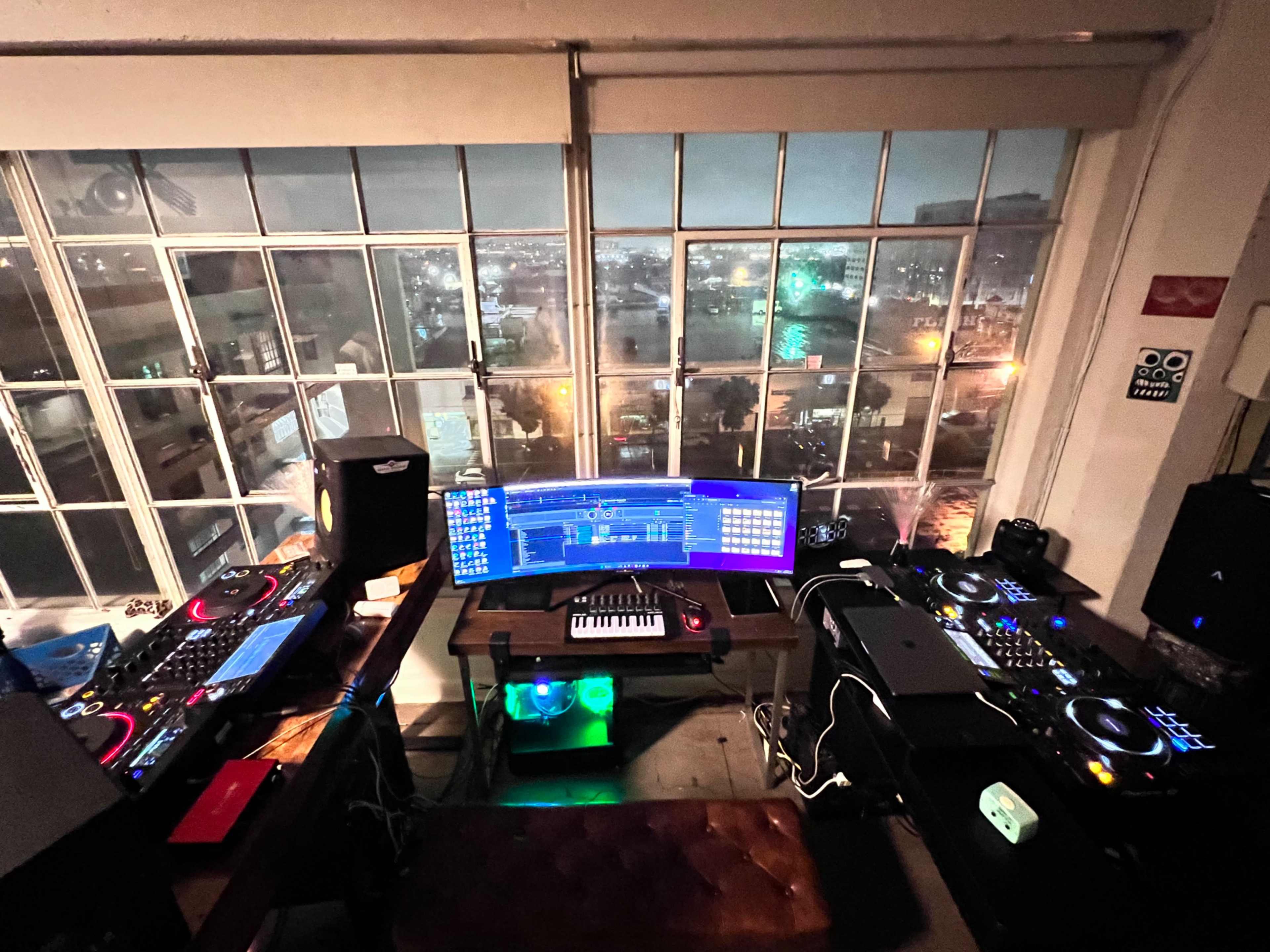 A dimly lit music studio features a large window overlooking a cityscape, with DJ equipment and a keyboard arranged on two desks.