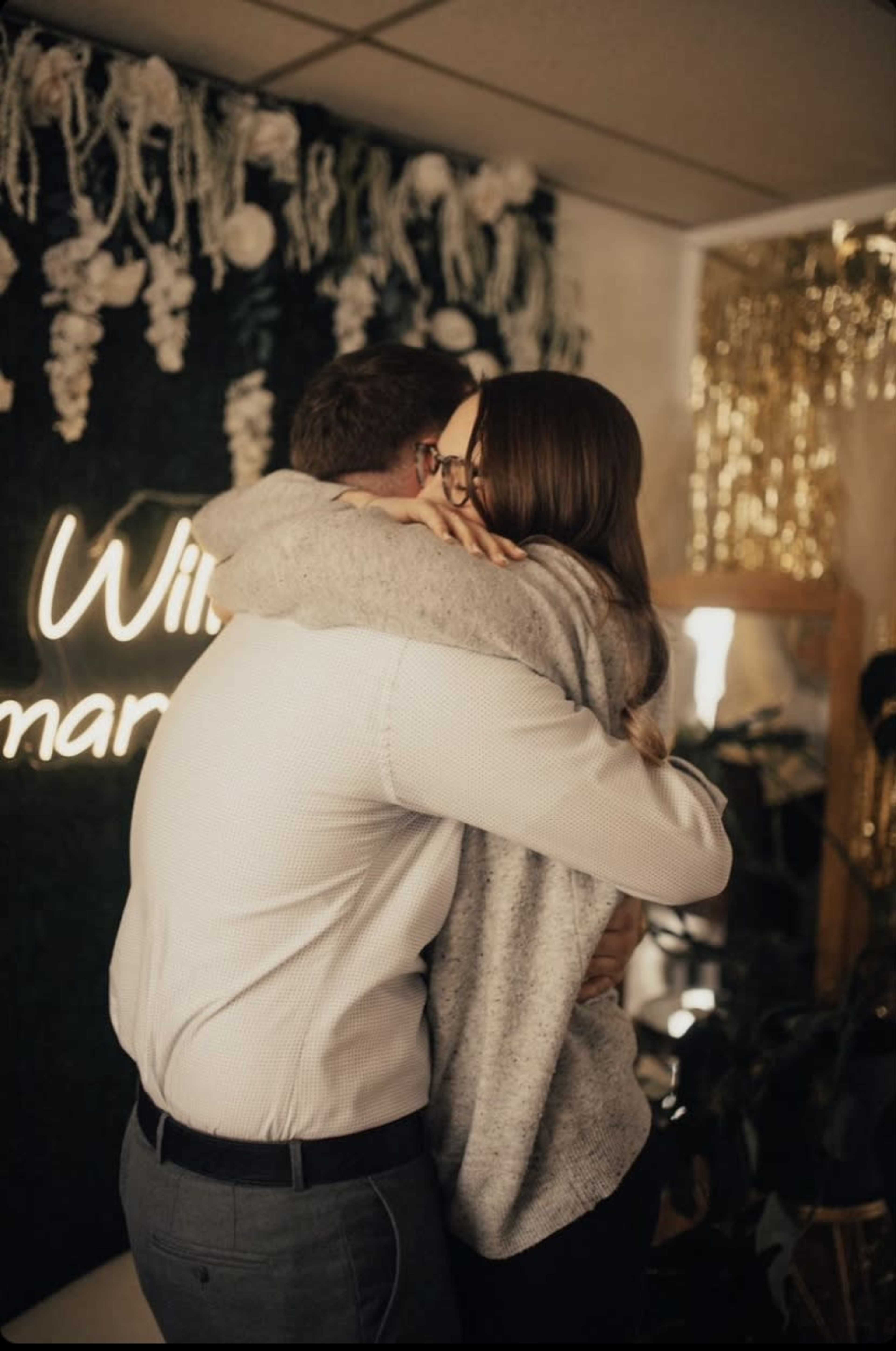 A man and a woman are embracing in a warmly decorated indoor space with a wall of greenery and decorations in the background.