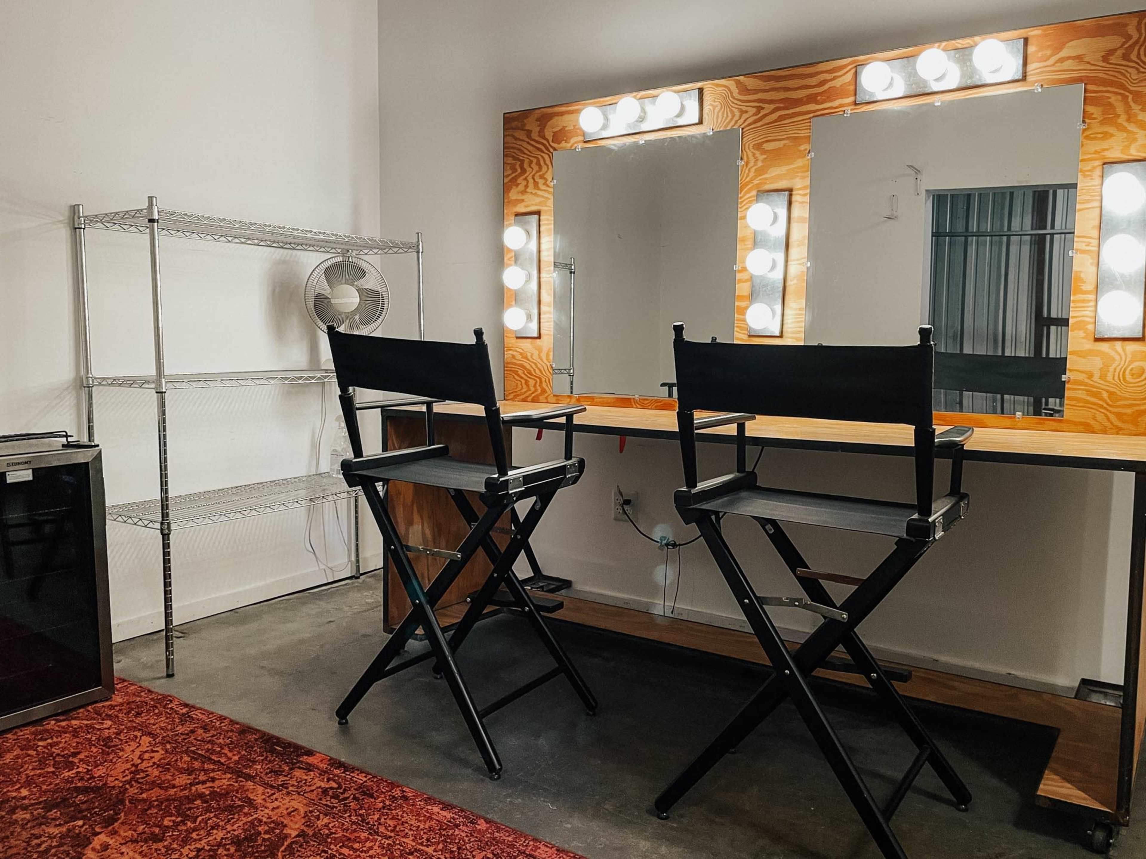 The image shows a well-lit makeup room featuring a large mirror with bright lights, two black director's chairs, a wooden table, and a metal shelving unit.
