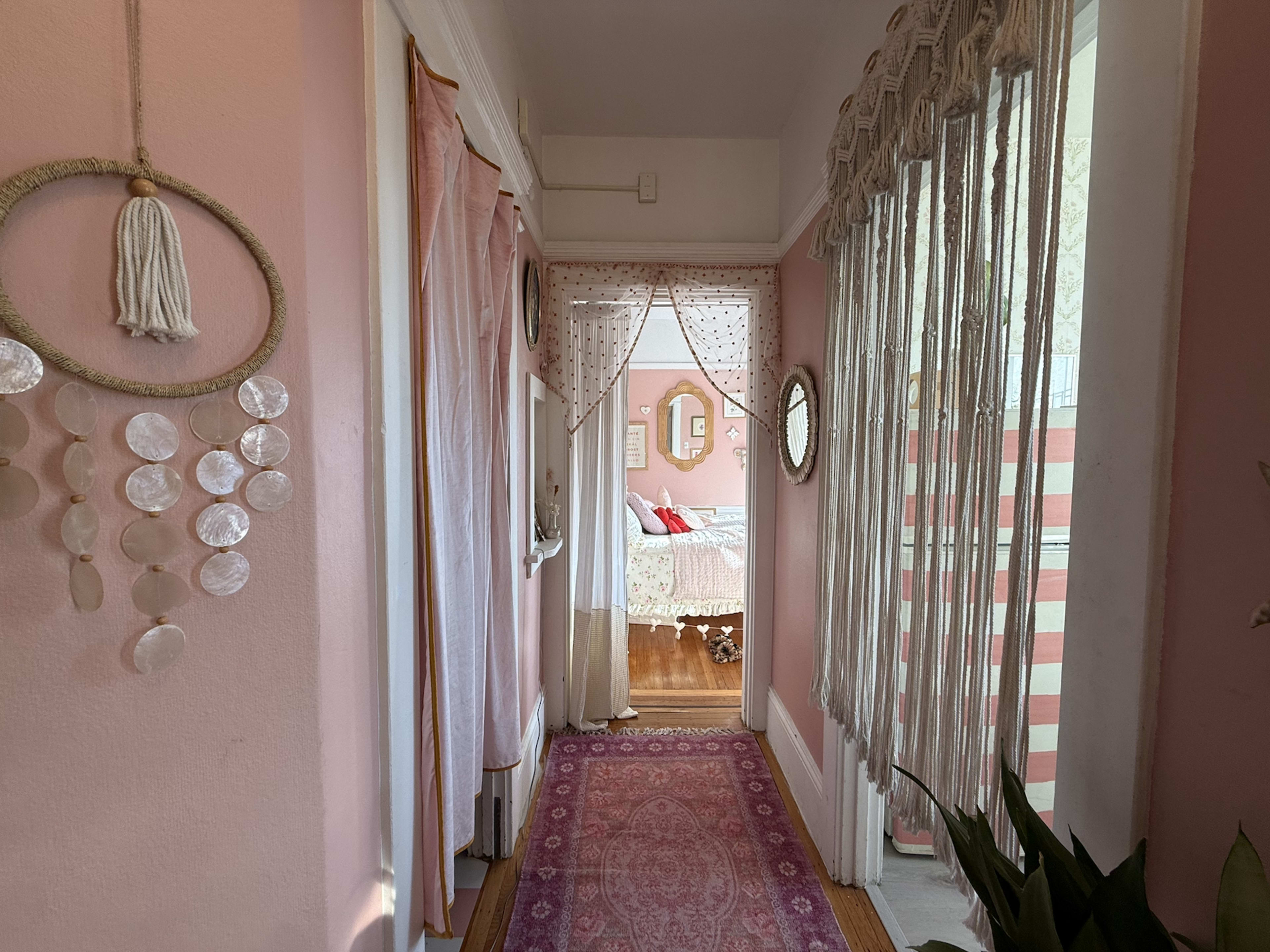 A pink hallway features draped curtains, decorative wall hangings, and a patterned rug leading to a visible room in the background.
