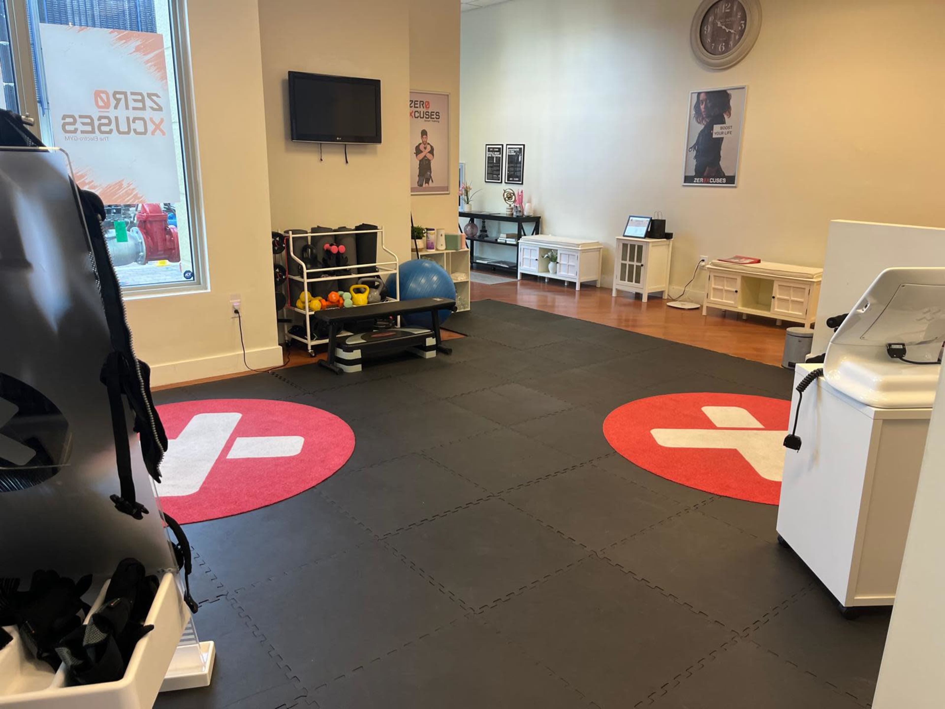 The image shows a fitness studio with exercise equipment, black rubber flooring, and a large red circle with a white cross symbol in two areas of the space.