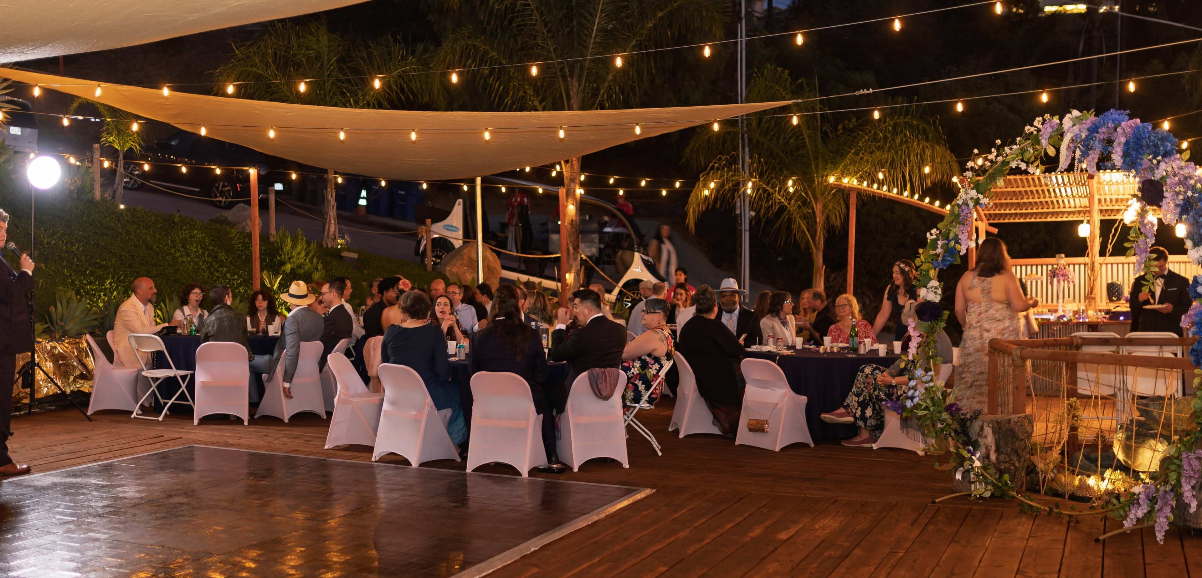 A decorated outdoor venue features guests seated at tables under string lights, with a floral backdrop and a speaker at a podium.