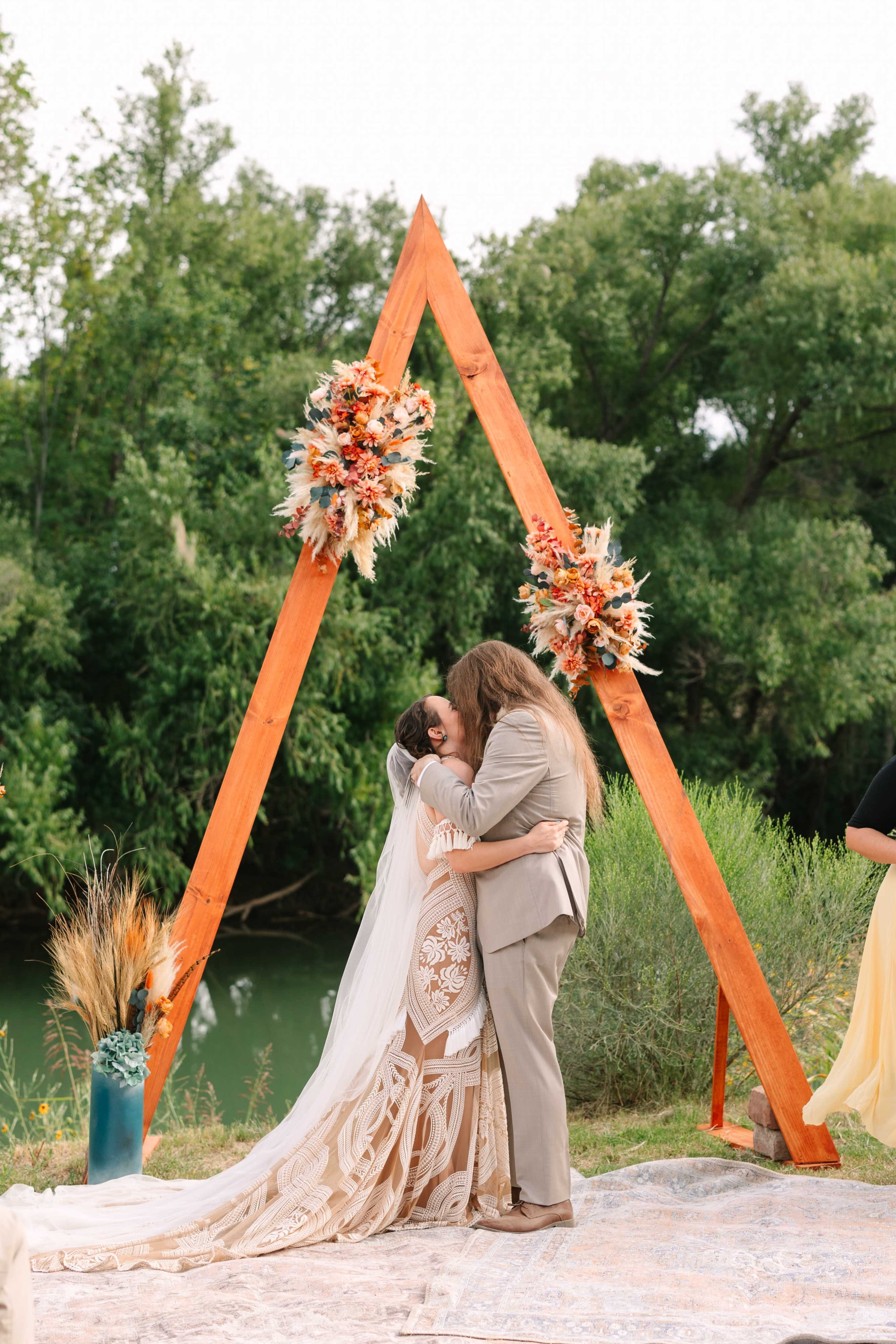 Two brides embrace under a wooden triangular arch adorned with flowers near a body of water.