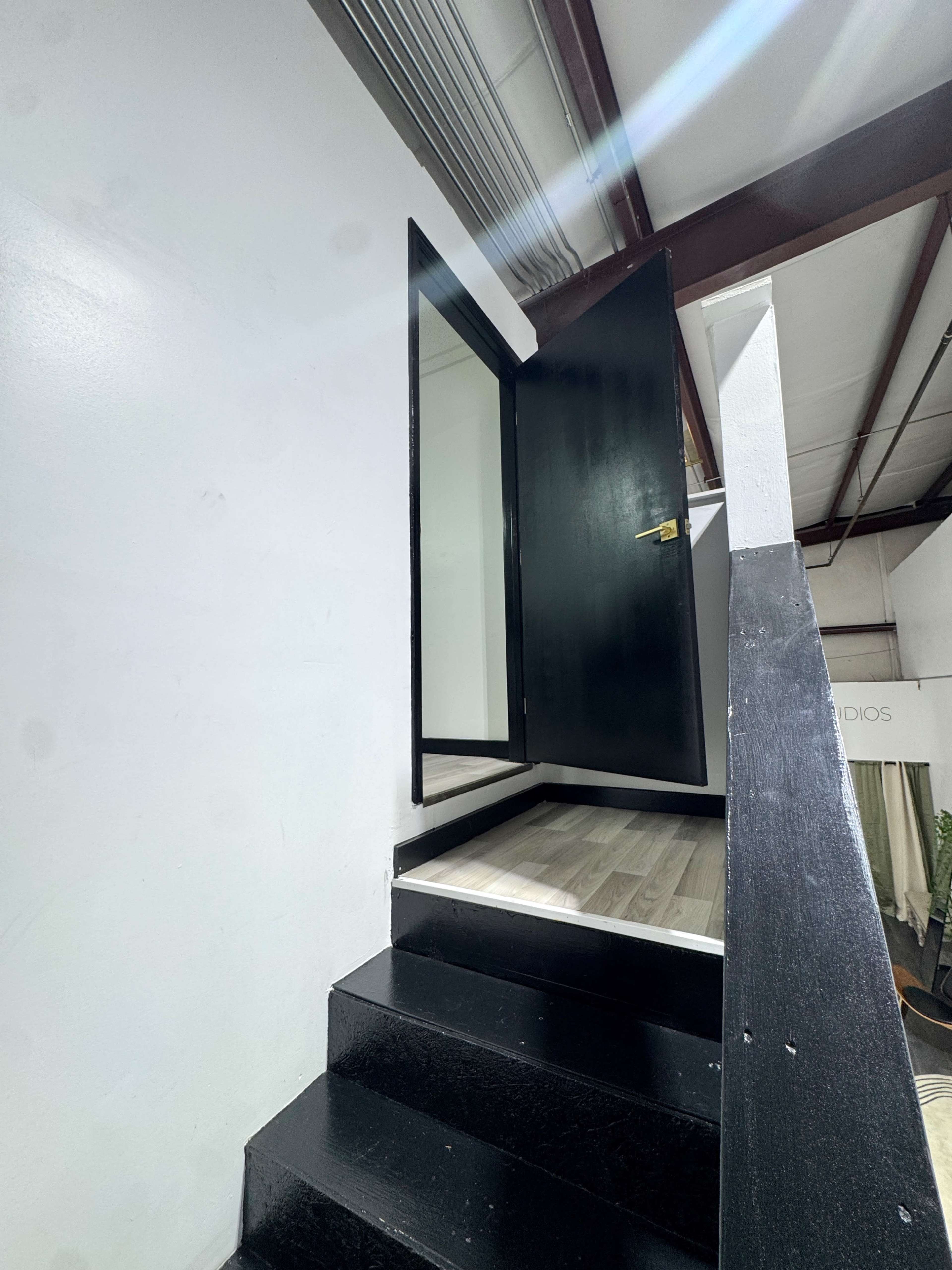 A black door is open at the top of a staircase, leading to a bright space with white walls and a wooden floor.