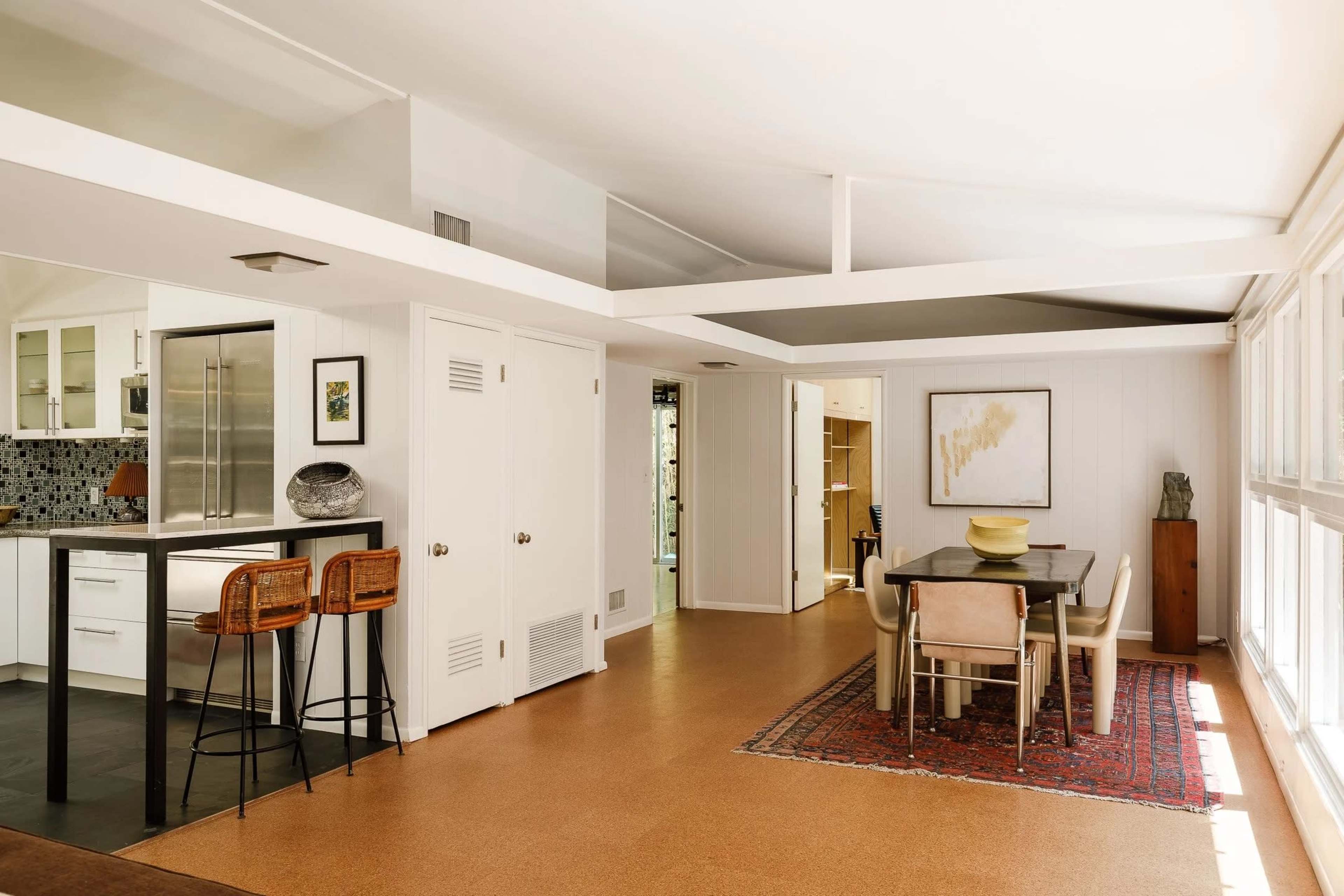 The image shows a modern interior space featuring a dining area with a table and chairs, an open kitchen with a bar counter, and natural light illuminating the room through large windows.