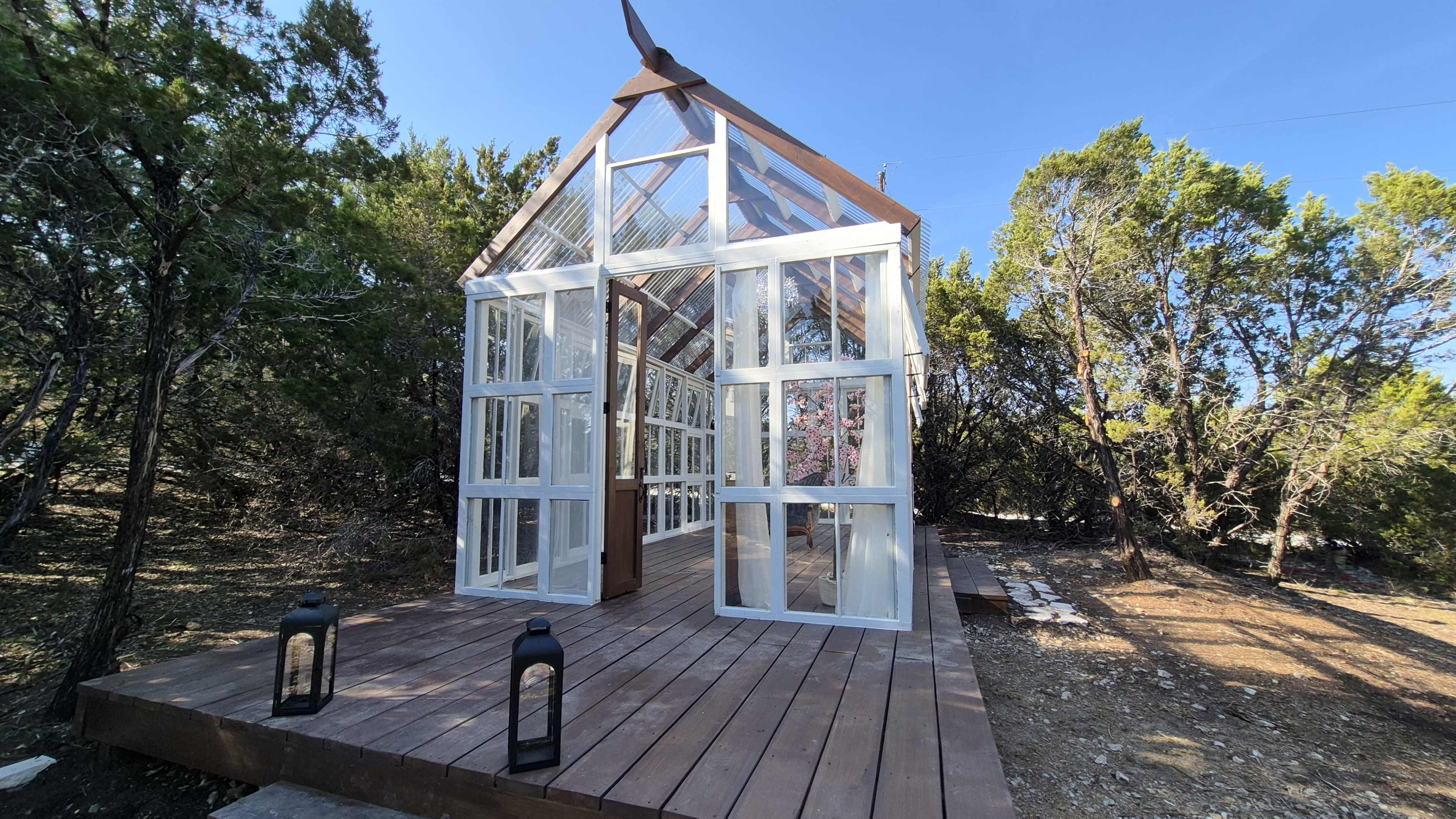 The image shows a glass-enclosed greenhouse with wooden flooring, situated among trees in a natural outdoor setting.