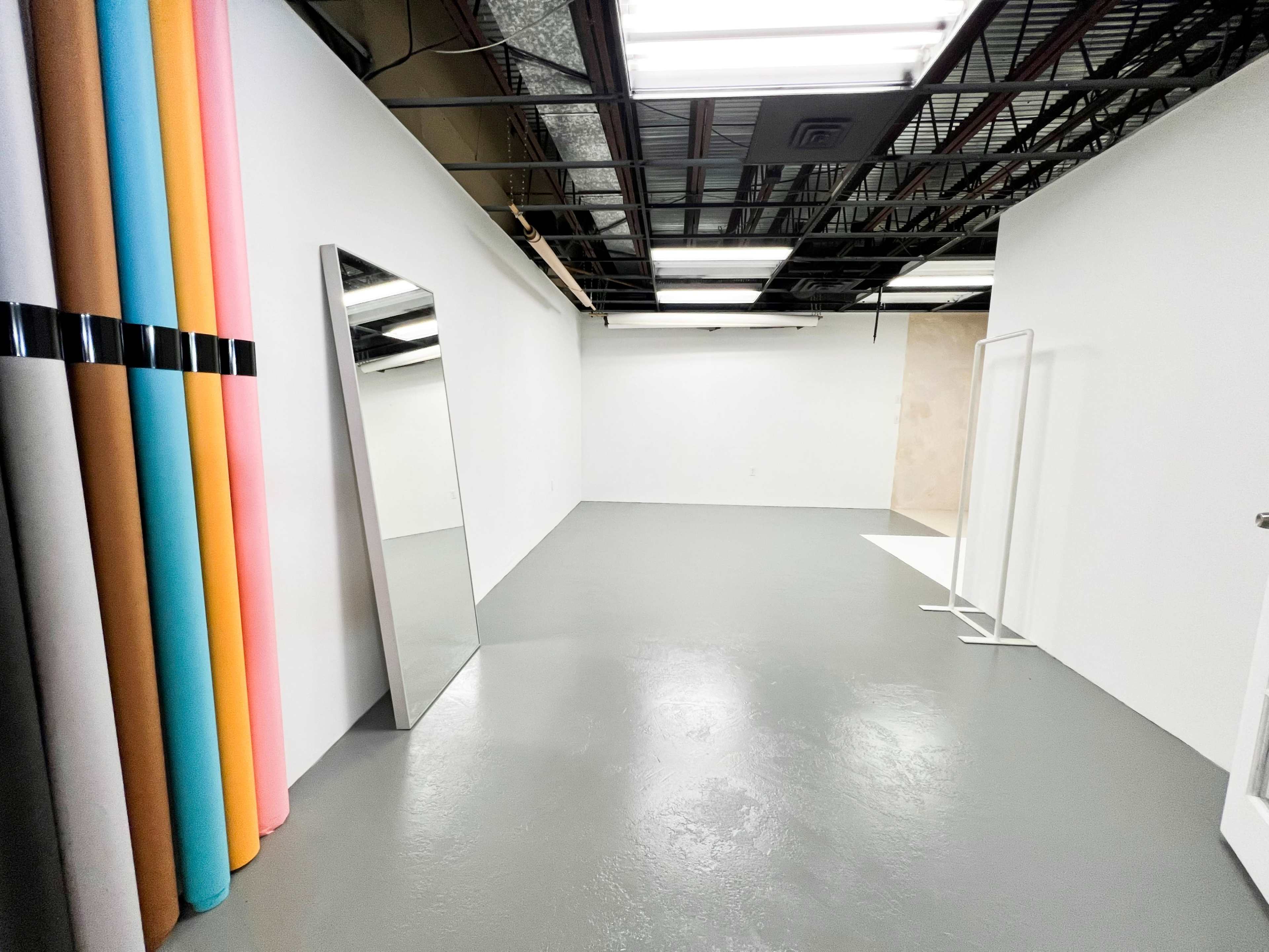 The image shows a spacious, empty room with a large mirror on one wall, a clothing rack, and rolls of colored paper leaning against another wall.