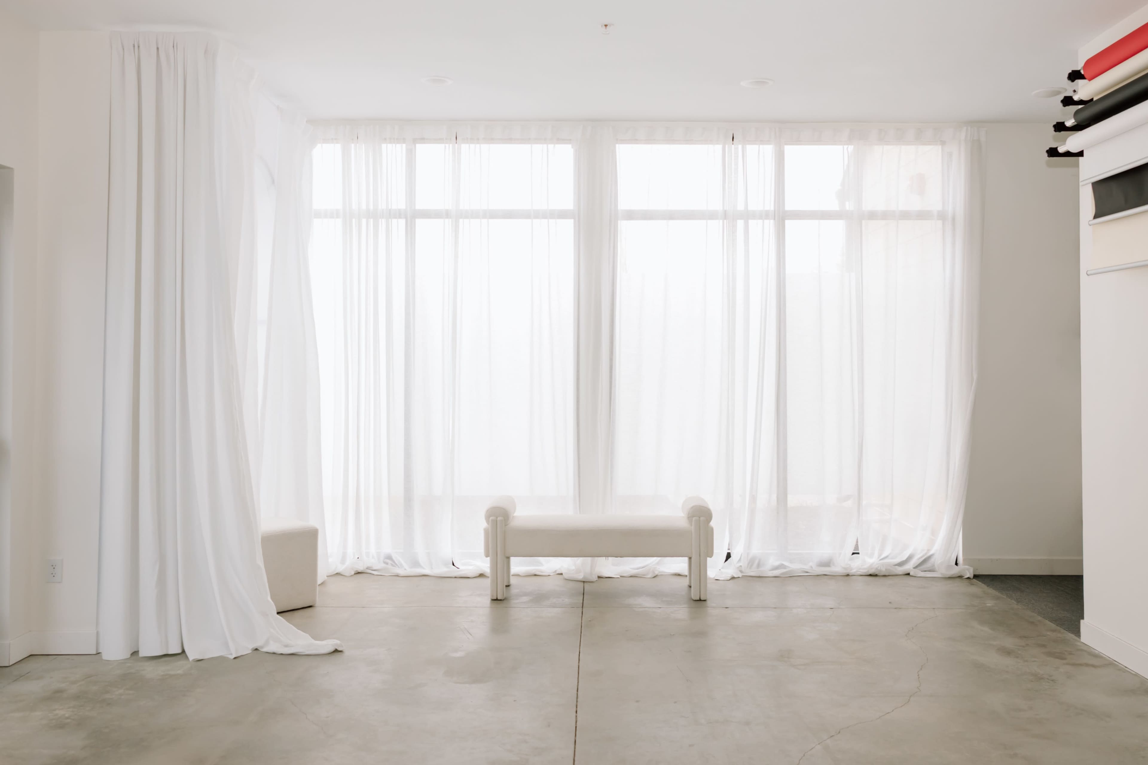 The image shows a bright, minimalistic interior with large sheer curtains, a white bench, and concrete flooring.