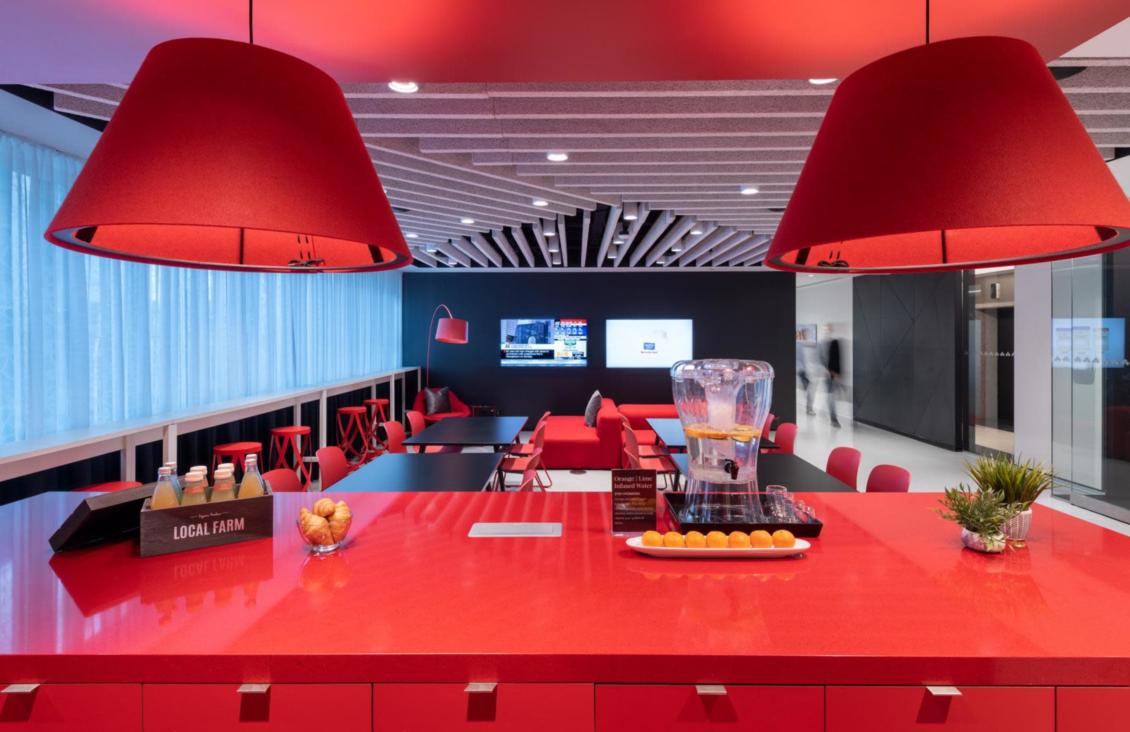 A modern red-themed lounge area with a glossy countertop, large pendant lamps, and seating arrangements against a black wall featuring a television screen.
