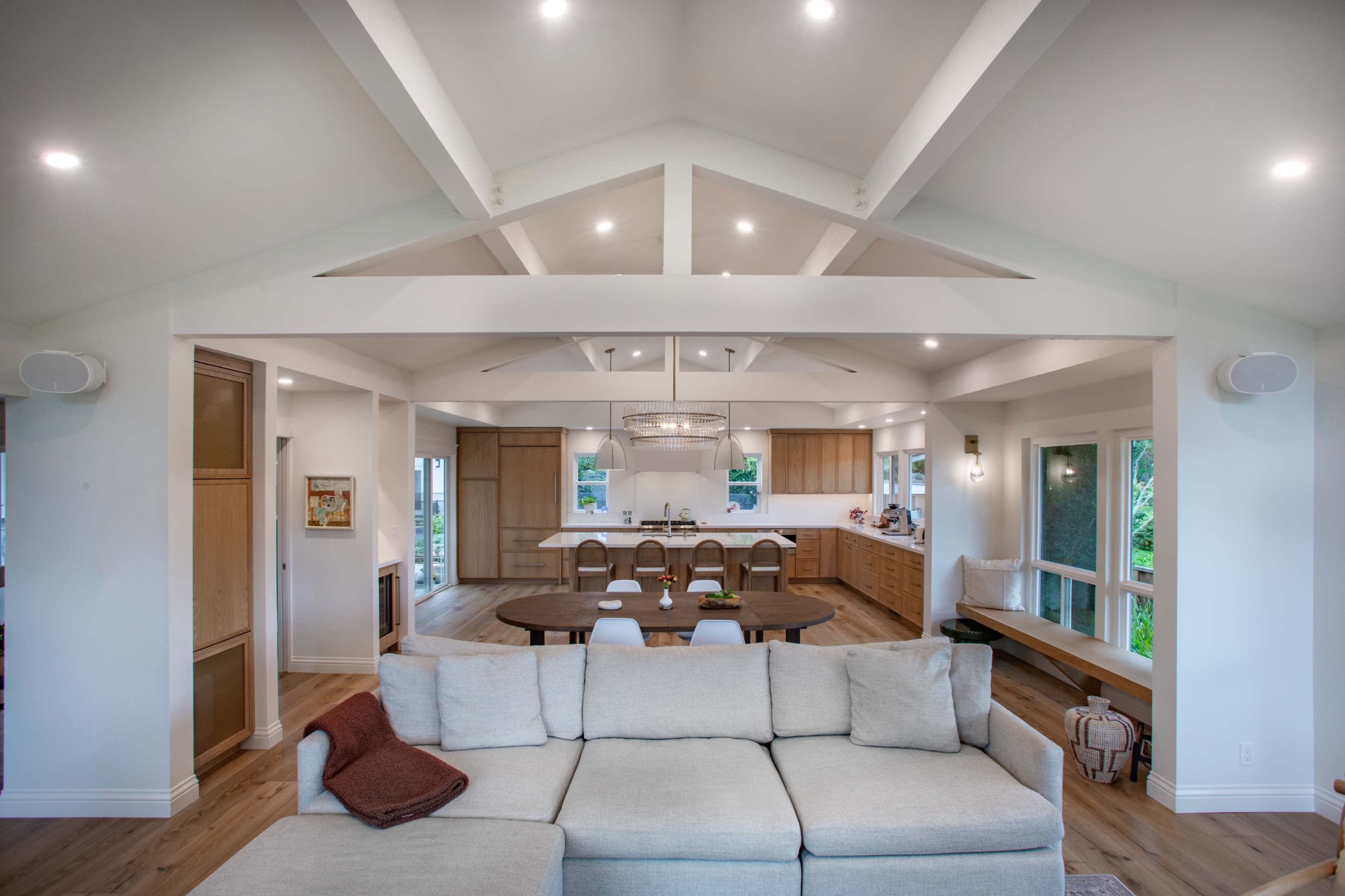 A modern, open-concept living space features a light-colored sofa in the foreground with a kitchen and dining area visible in the background, characterized by wooden cabinetry and high ceilings with exposed beams.